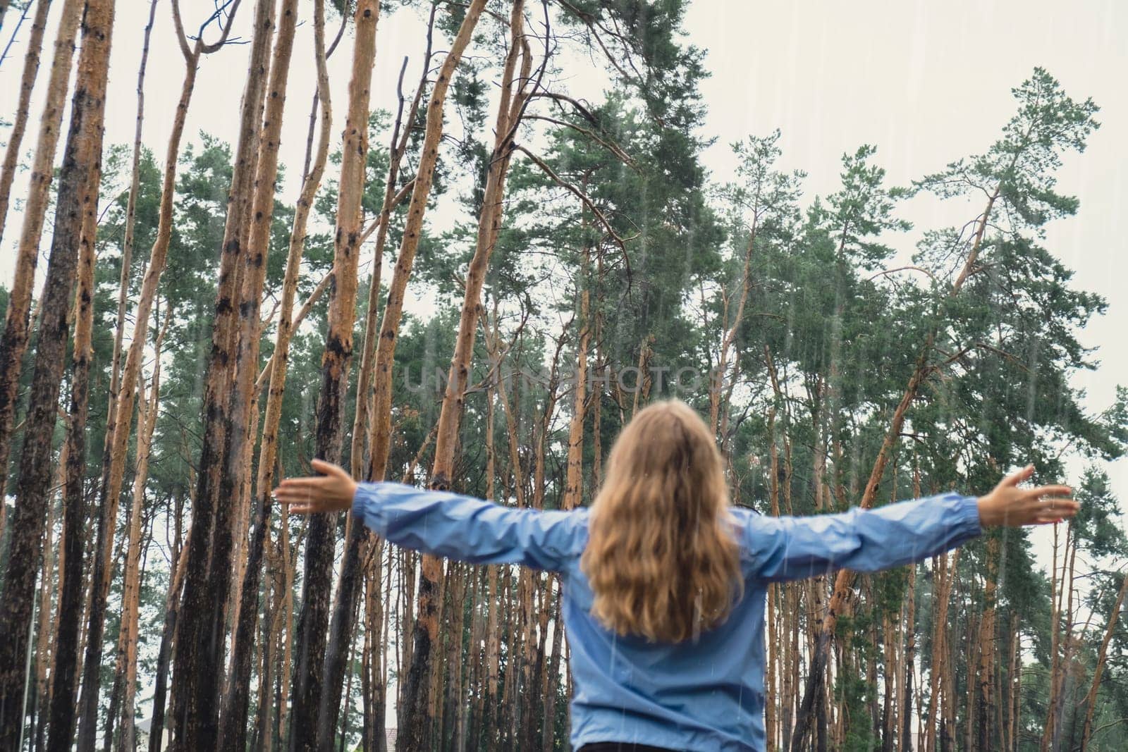Out of focus Back view young woman walk in blue raincoat enjoying the woods in park. Open arms outdoors in rainy weather forecast. Tourist rest and feel freedom. Autumn season