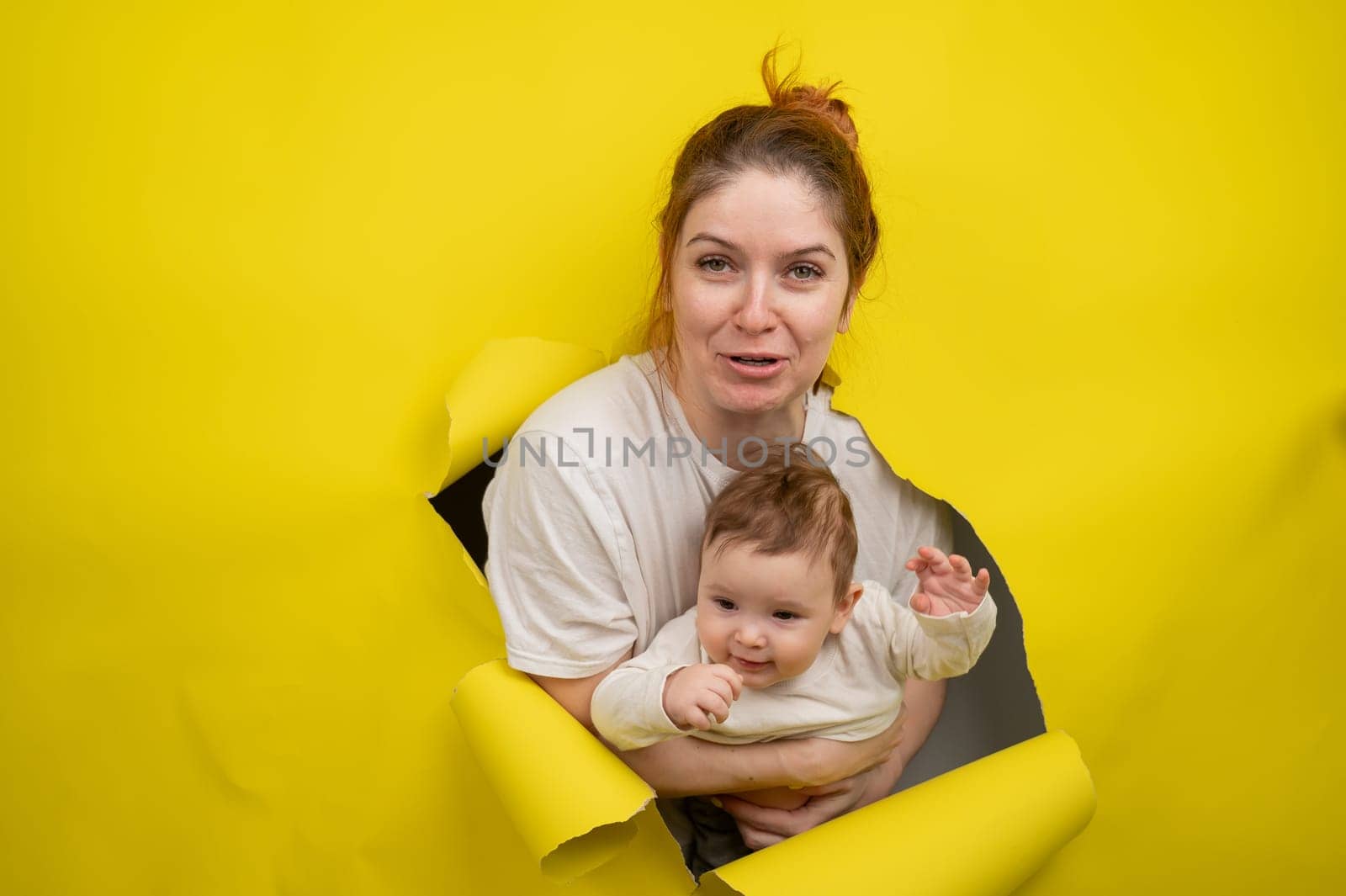 Cheerful Caucasian woman with little son rips and leans out through yellow cardboard background