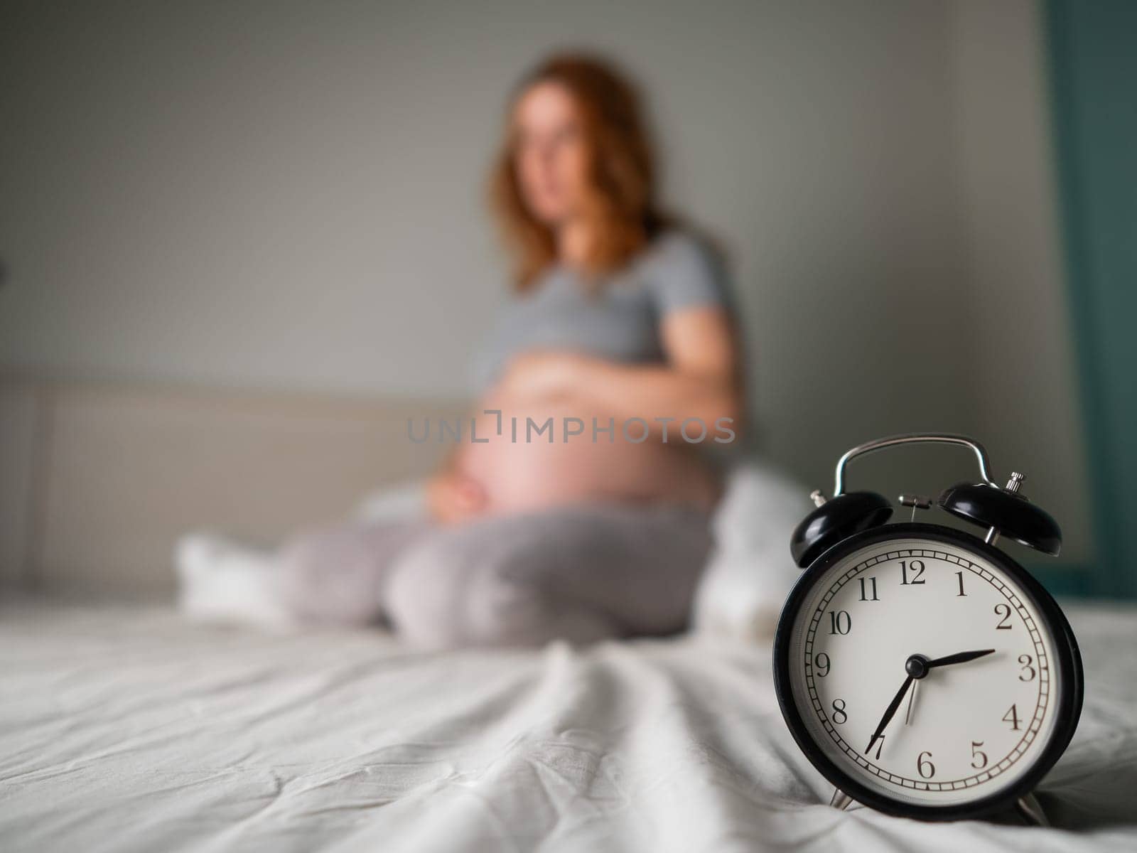 Caucasian pregnant woman sits on the bed and suffers from insomnia. Alarm clock in the foreground