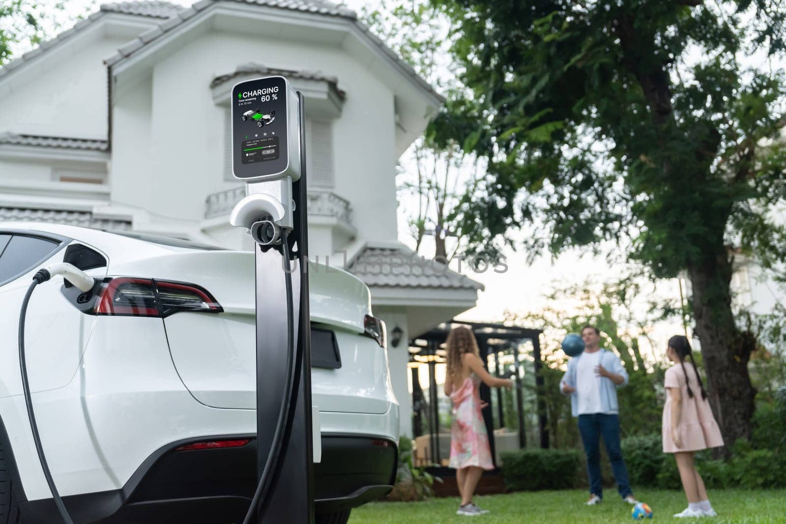 Focus electric vehicle recharge from home charging station on blur background of happy and playful family playing together. EV car using alternative and sustainable energy for better future.Synchronos