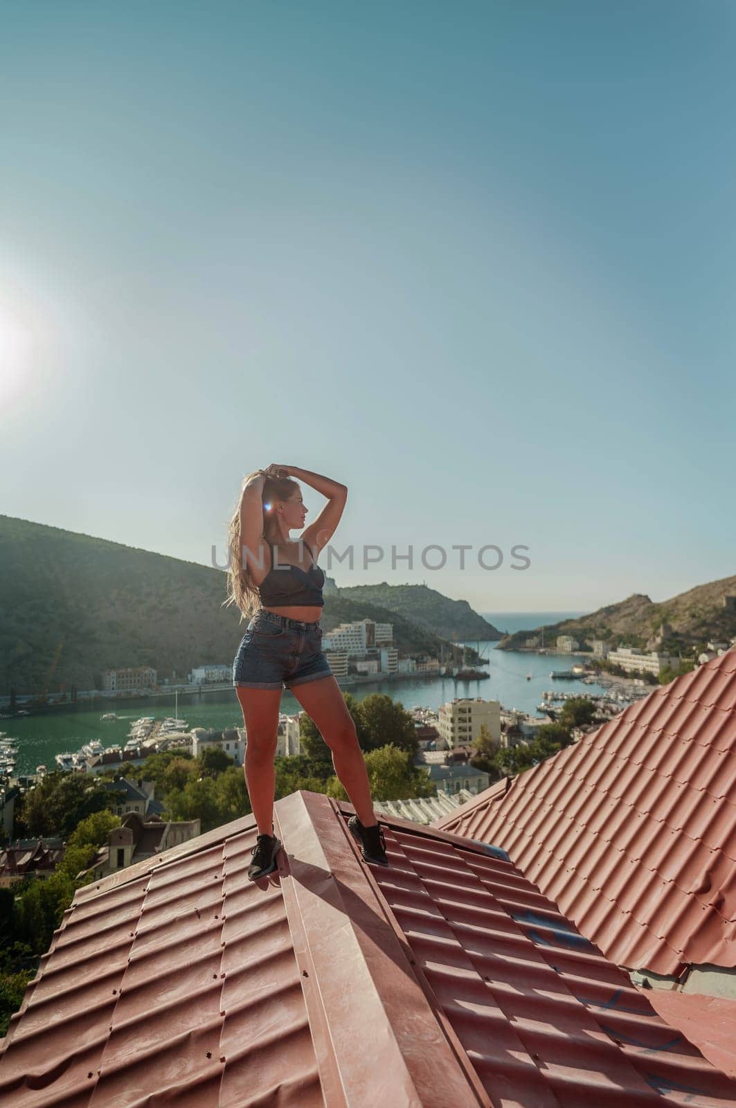 Woman standing on rooftop, enjoys town view and sea mountains. Peaceful rooftop relaxation. Below her, there is a town with several boats visible in the water. Rooftop vantage point