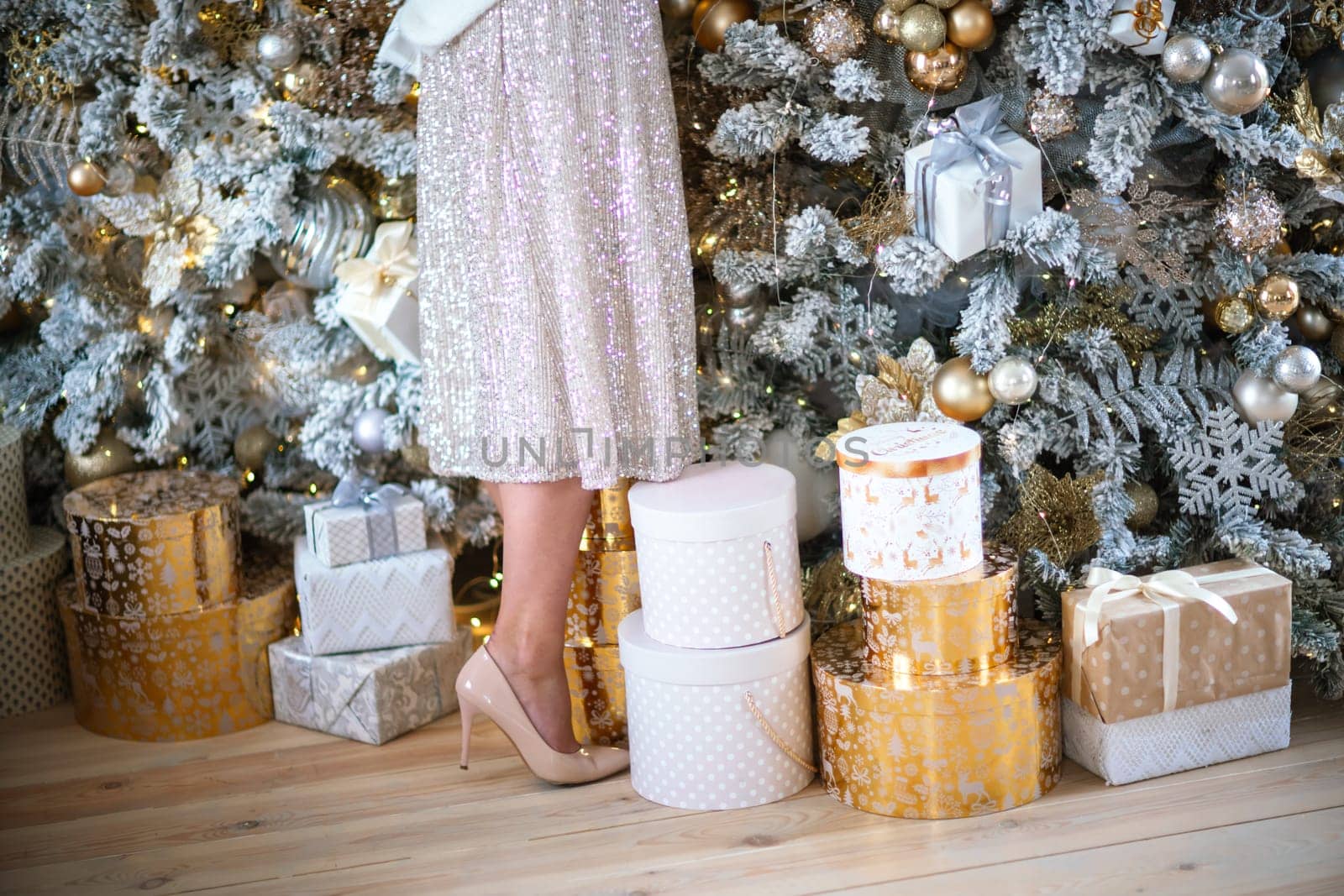 Women's legs. Cropped close-up photo of healthy beautiful elegant female legs in beige high-heeled shoes, a woman stands on the background of a Christmas tree with gifts by Matiunina