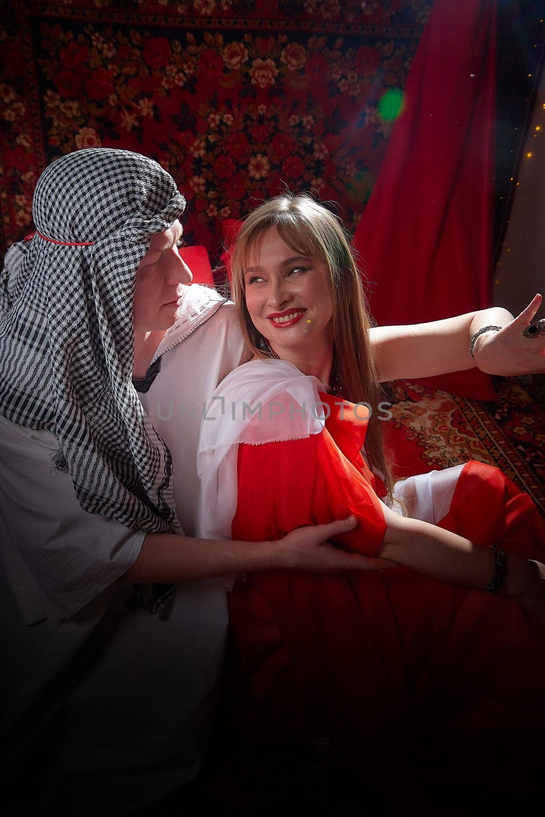 Portrait of young arabian muslim couple in traditional clothes in cozy red room. Fhoto shoot in easten style with male and female model like in harem with a sultan and an odalisque. Partial focus