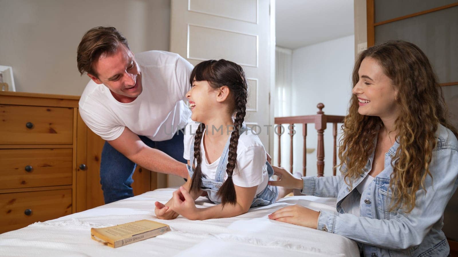 Happy modern family wakes up their little girl on the weekend with playful tickle expressing their love and affection for their young daughter, laughing and smiling together. Synchronos