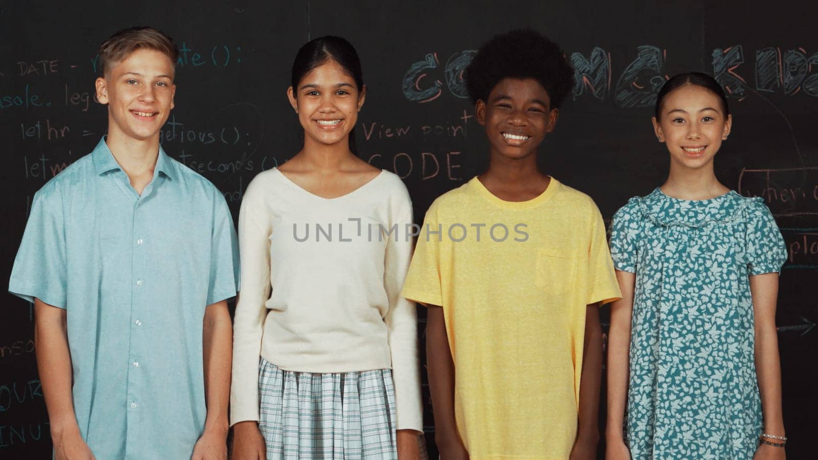 Group of smart multicultural teenager looking at camera at blackboard. Happy diverse children standing in STEM technology class with engineering code, prompt or planning program idea. Edification.