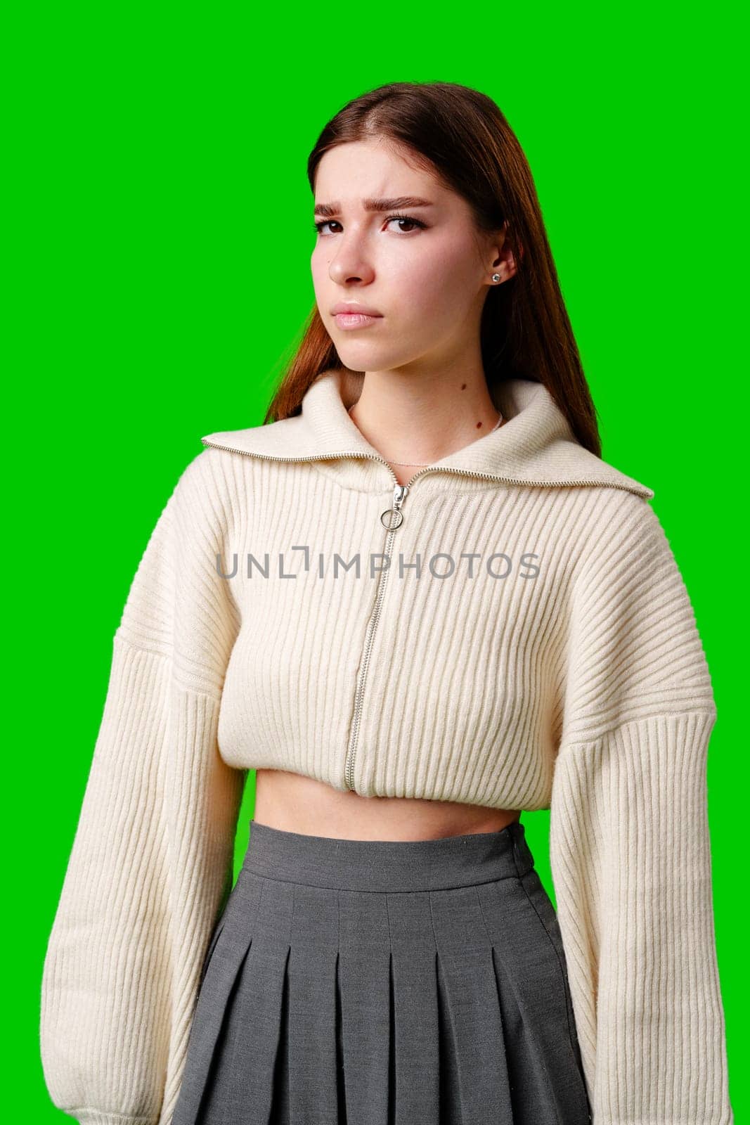 A young woman stands confidently against a vivid green background, dressed in a casual yet fashionable white cropped hoodie and a gray pleated skirt. Her expression is serene with a hint of contemplation, and her body language exudes a relaxed poise.