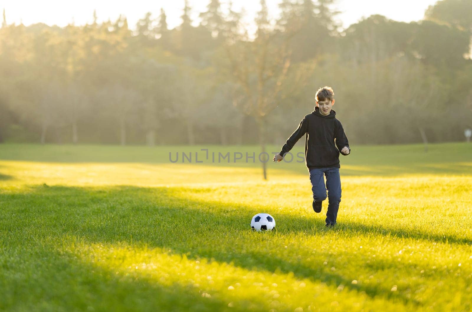A boy is playing soccer in a field. The grass is green and the sky is blue. The boy is wearing a black jacket and blue jeans