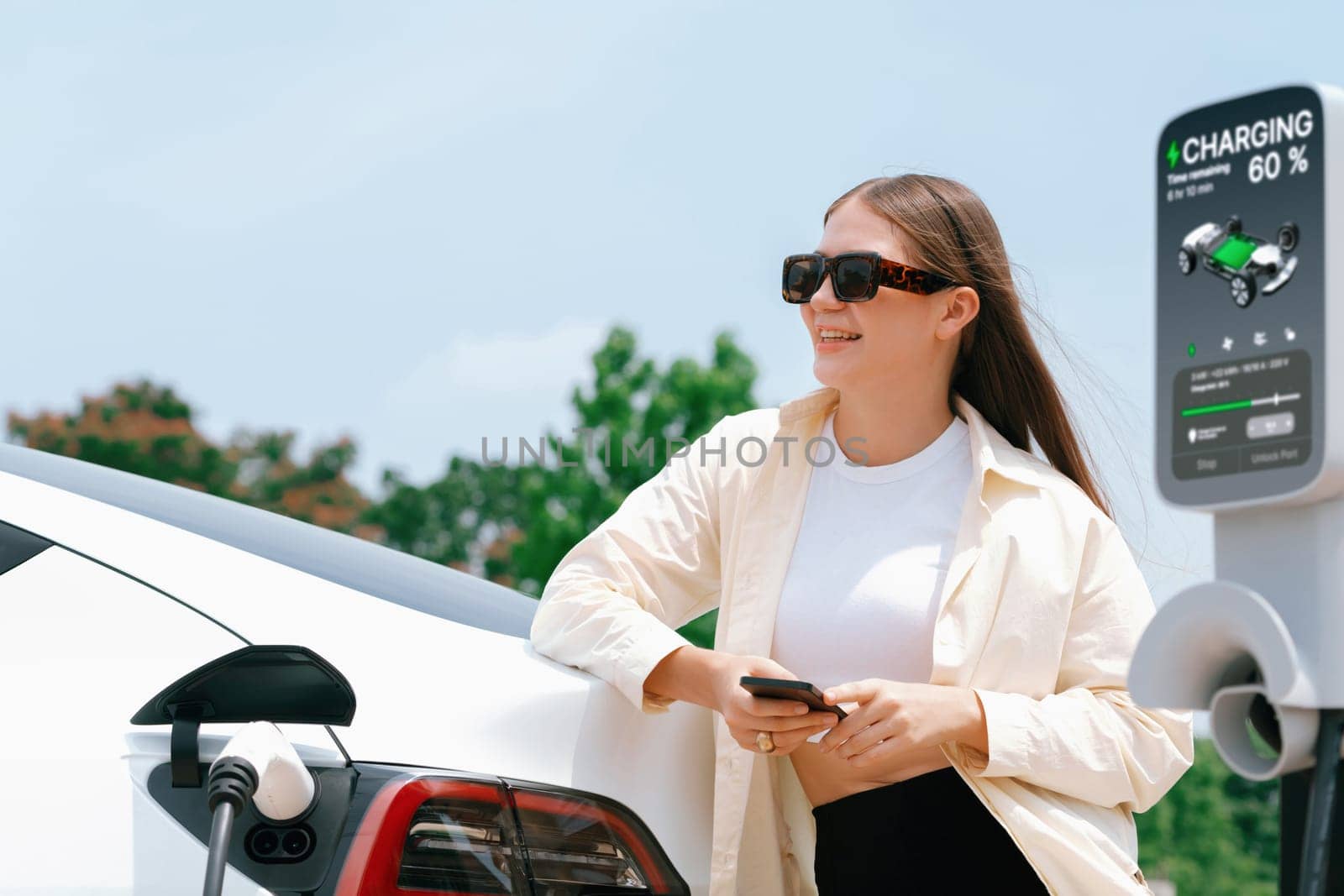 Young woman use smartphone to pay for electricity at public EV car charging station green city park. Modern environmental and sustainable urban lifestyle with EV vehicle. Expedient