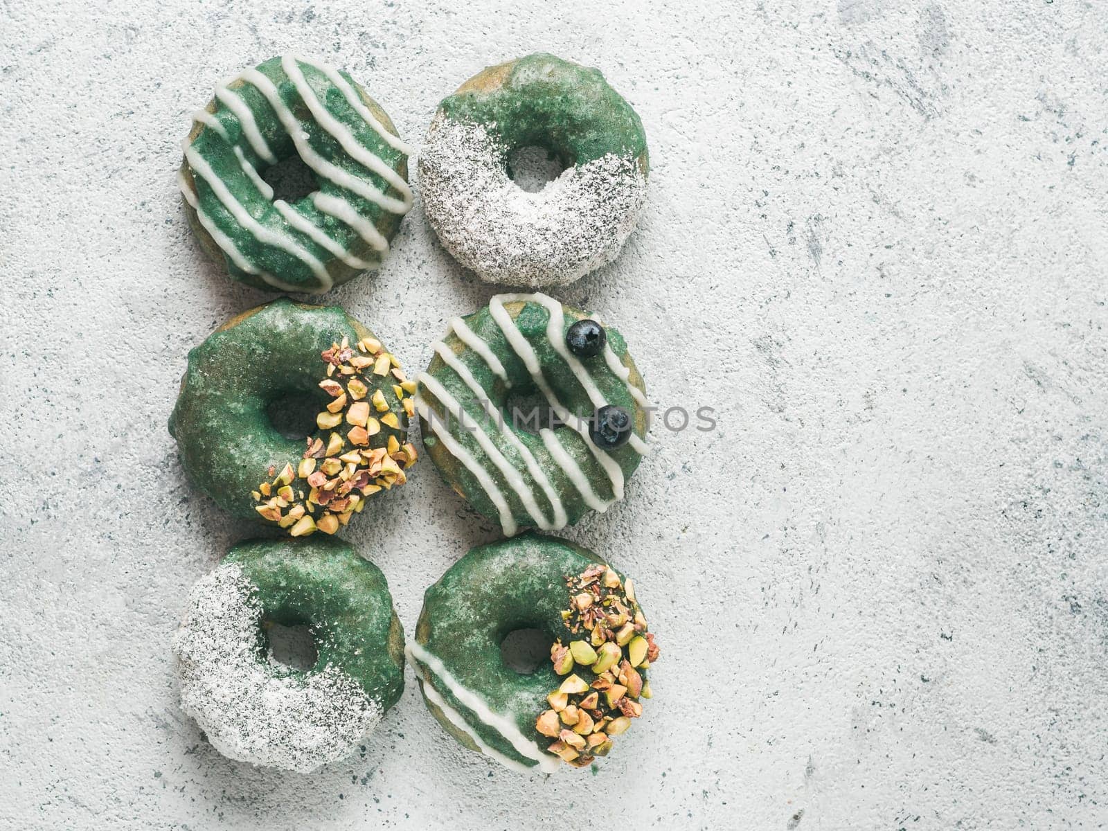 Vegan doughnuts with chia seeds topped with healthy spirulina glaze with pistachio, desiccated coconut and blueberry. Blue green spirulina donuts, on gray background, top view or flat lay
