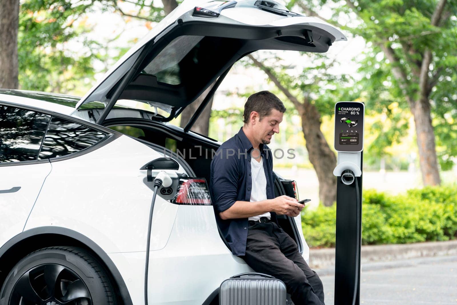 Man using smartphone online banking application to pay for electric car battery charging from EV charging station during vacation holiday road trip at national park or summer forest. Exalt