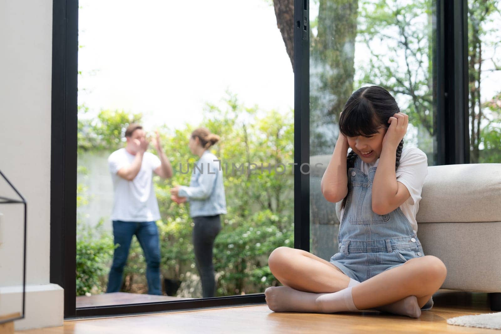 Stressed and unhappy girl huddle in corner, cover her ears with painful expression while her parent arguing in background. Domestic violence and traumatic childhood develop to depression. Synchronos