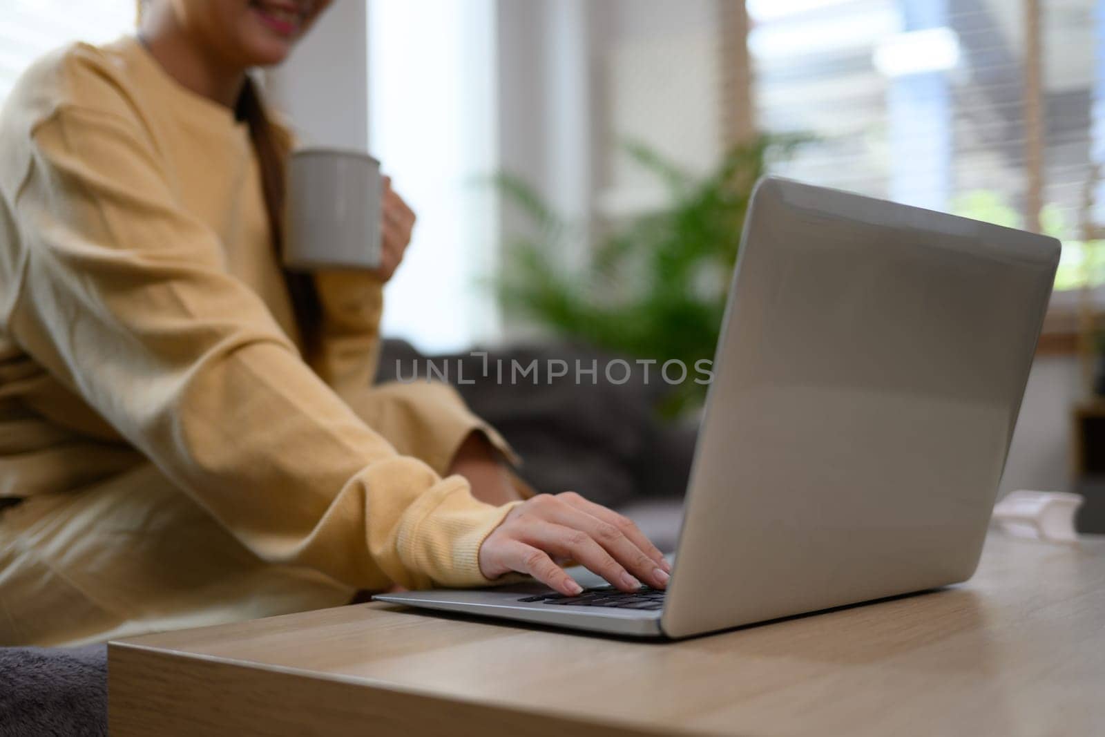 Young woman holding cup of coffee and using laptop in living room by prathanchorruangsak