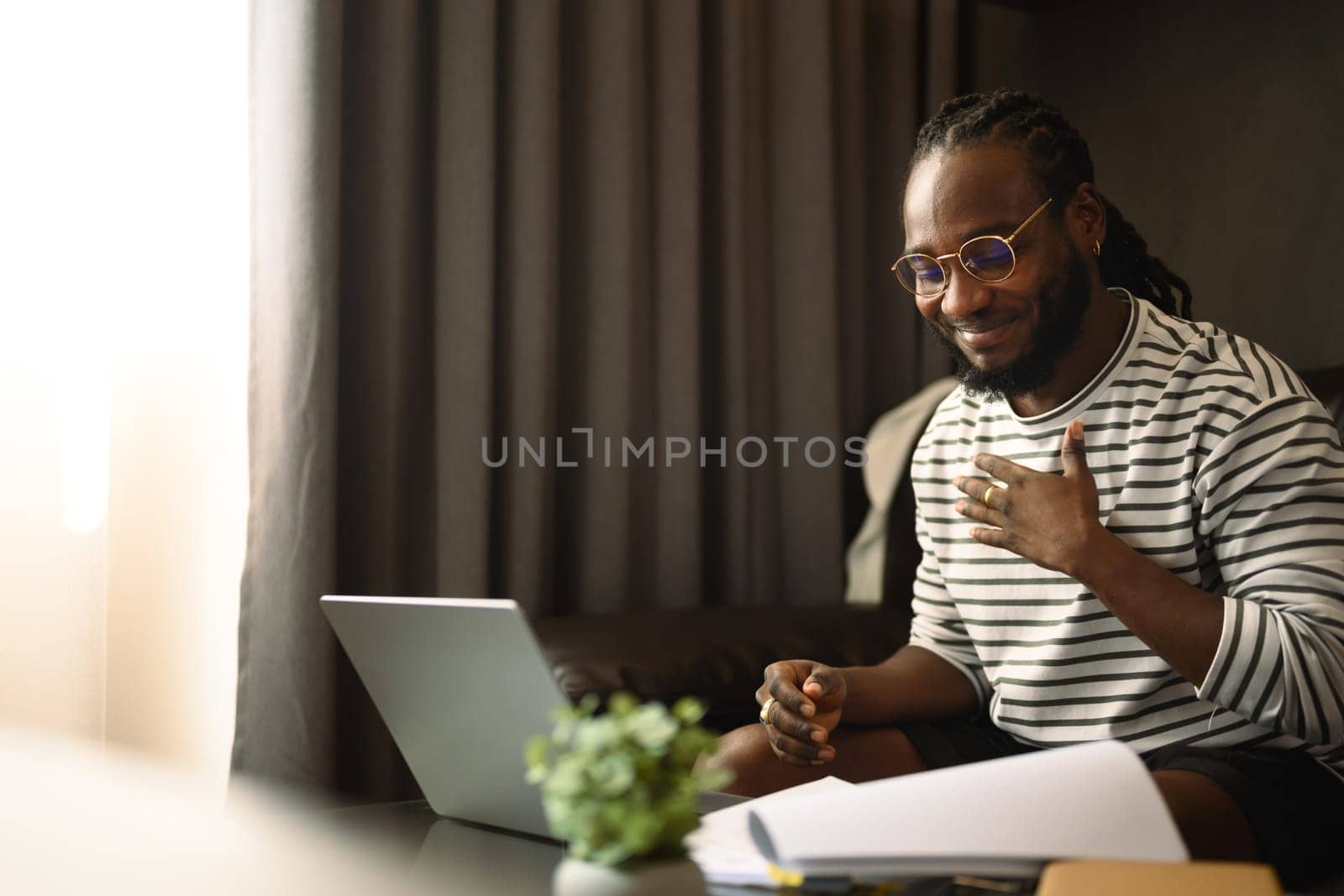 Smiling young African man freelancer having video call on laptop during working at home by prathanchorruangsak