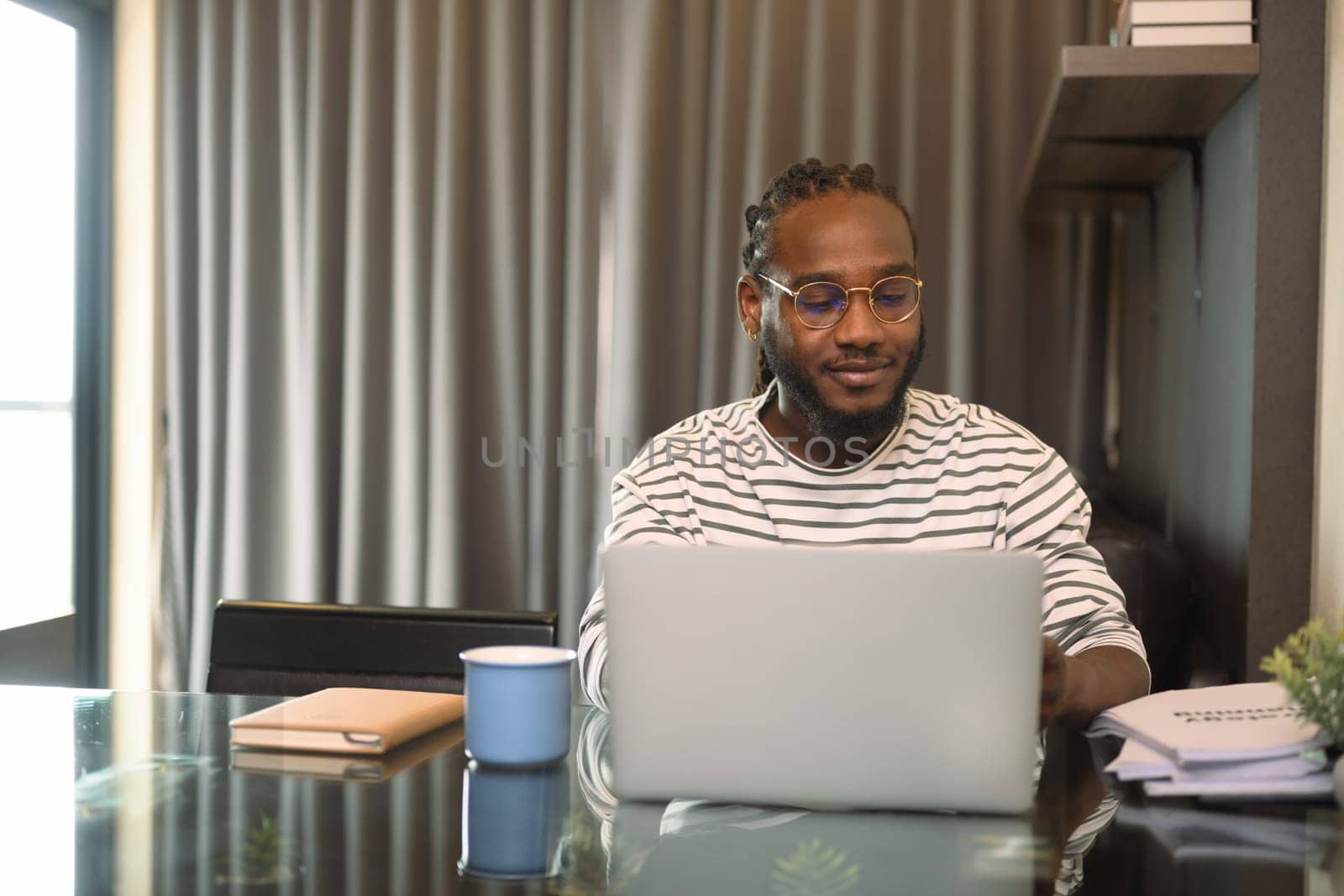 Smiling bearded African man working remotely from home using laptop on dinning table.