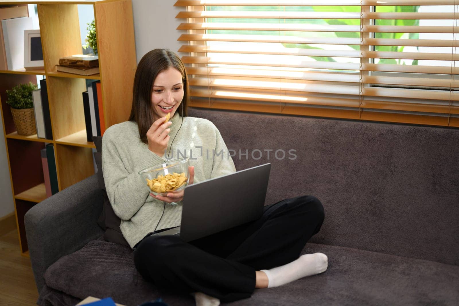 Relaxed young caucasian woman eating popcorn and watching movie on a laptop at home by prathanchorruangsak