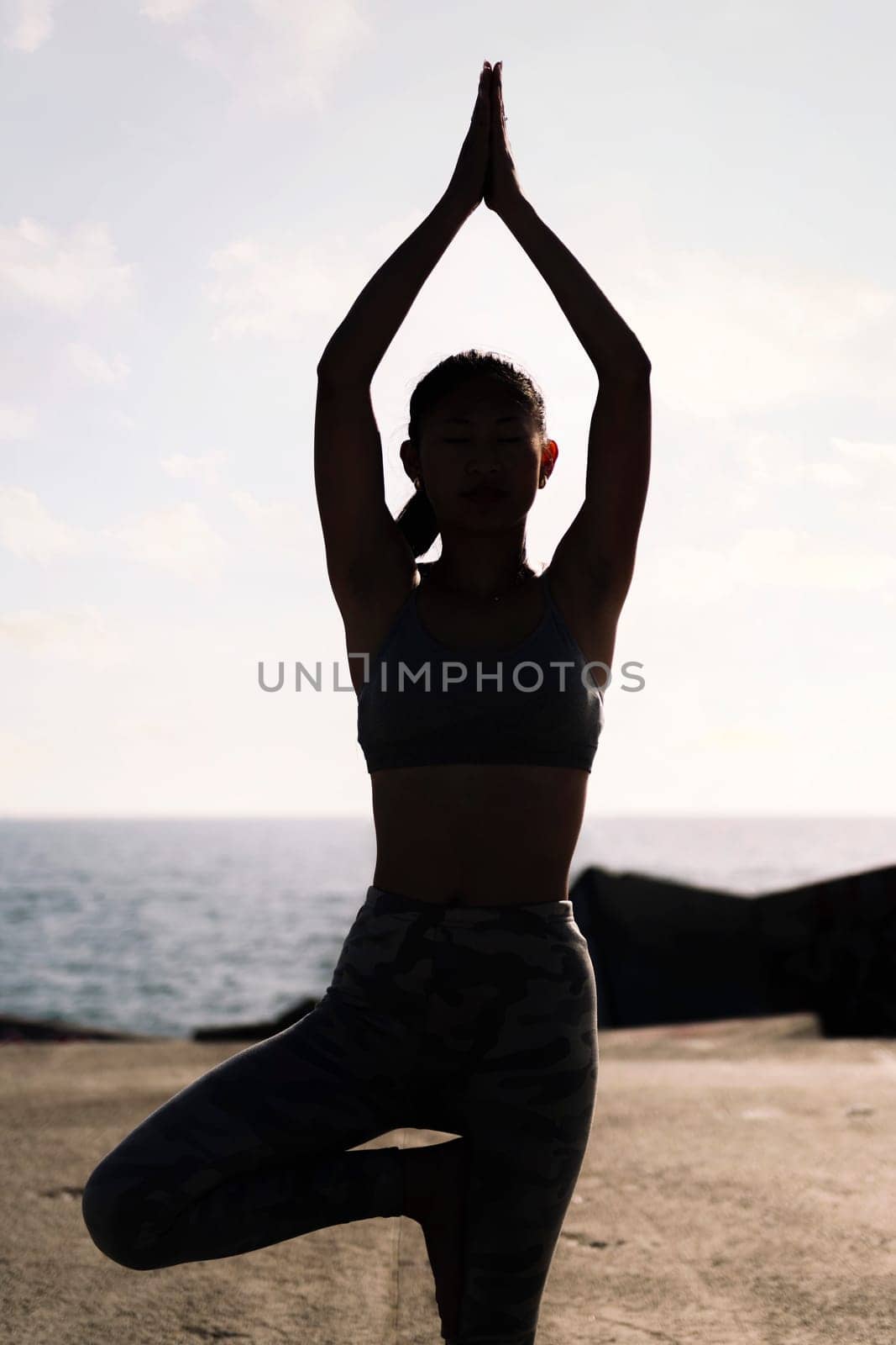 backlight of young woman in sportswear doing yoga tree position by the sea, concept of mental relaxation and healthy lifestyle