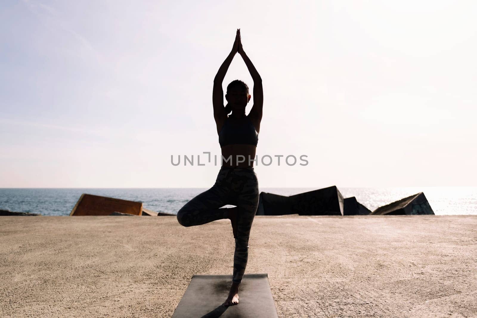 backlight of young woman in sportswear doing tree position on her yoga mat by the sea, concept of mental relaxation and healthy lifestyle