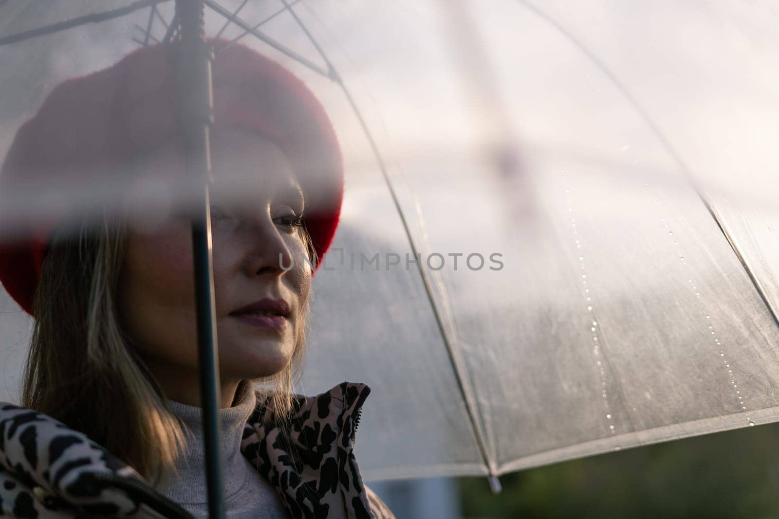 A woman wearing a red hat and a black and white jacket is holding a clear umbrella. The umbrella is open, and the woman is looking up at the sky. Concept of solitude and contemplation. by Matiunina