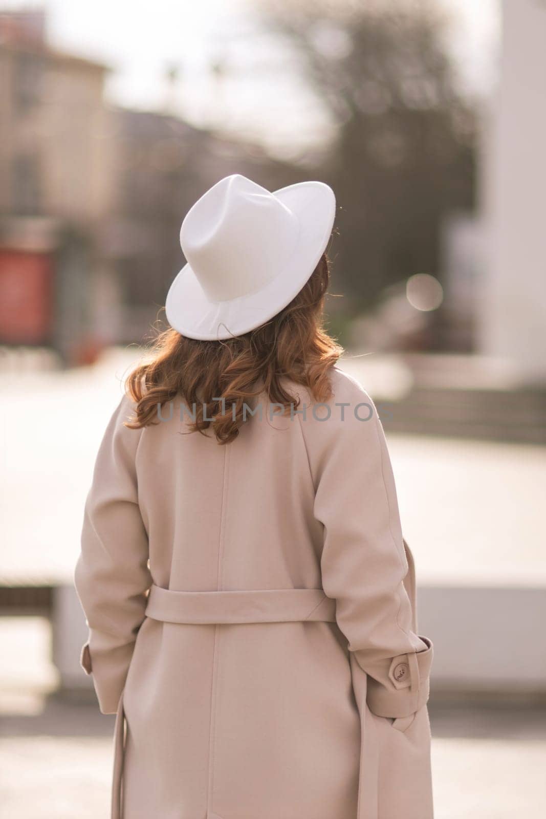 Back view of happy woman wearing hat and coat walking down street on sunny spring day. People, lifestyle, travel and vacations concept by Matiunina