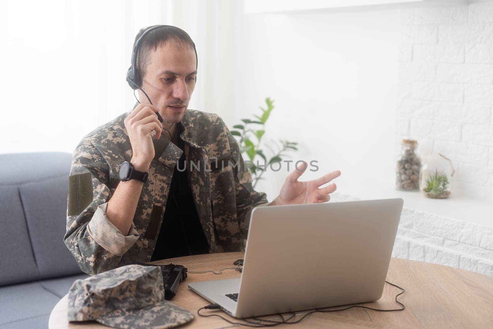 Soldier in office. Serious young military man typing on laptop while working in headquarters building. Caucasian man in camouflage uniform is sitting in bright room at table with notebook and cap. by Andelov13