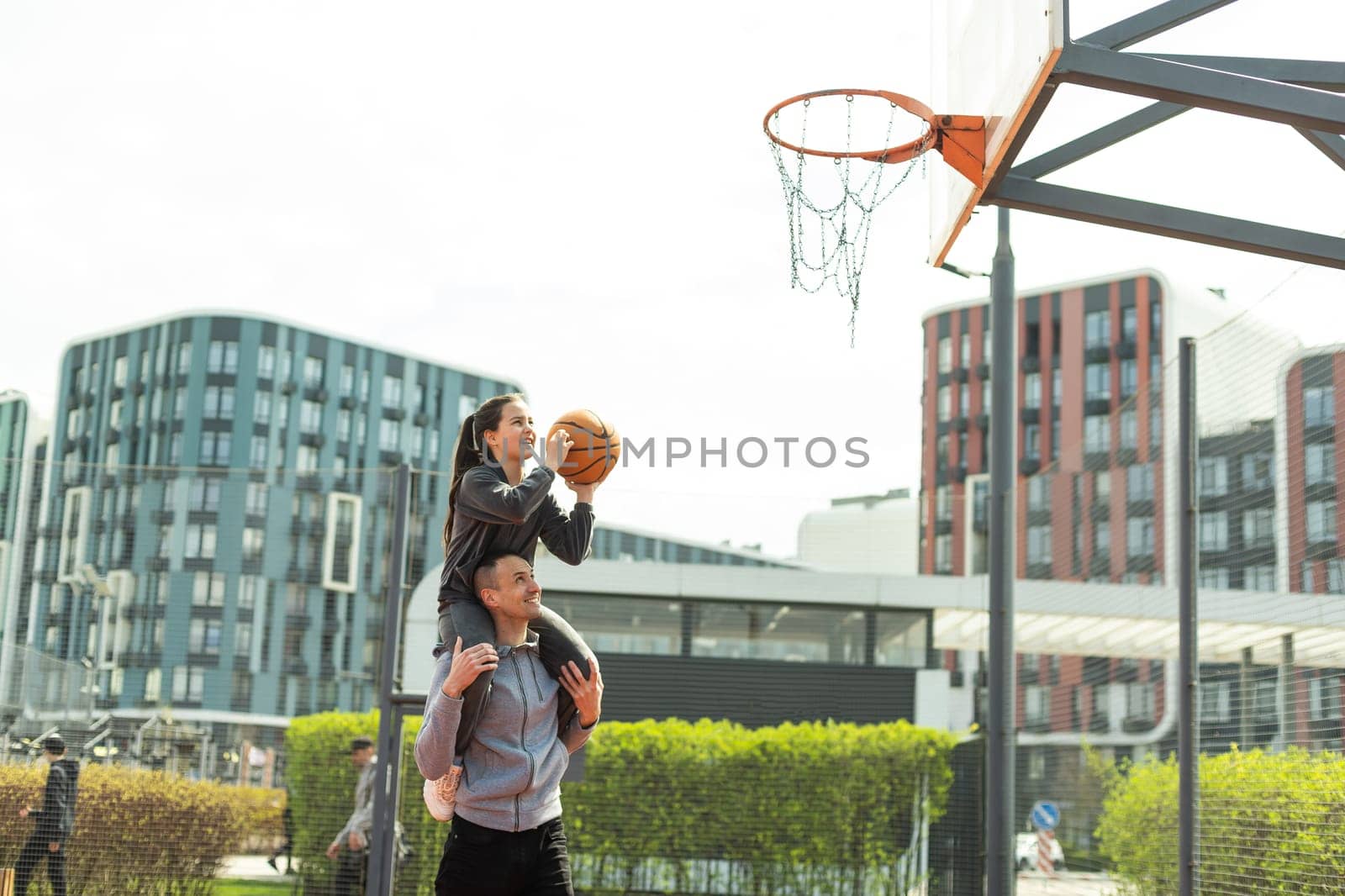 Father Carrying Daughter On Shoulders Helping Her To Put Basketball In Net