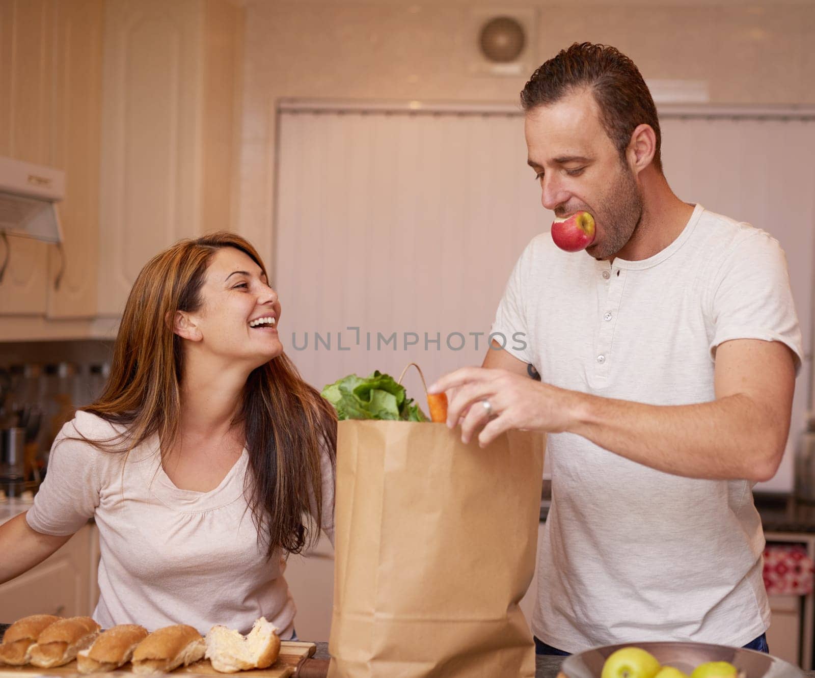 Brown bag, kitchen and couple with groceries, vegetables and funny with conversation and chatting. Healthy meal, food and ingredients with fruit and wellness with humor, home and laughing for joke.