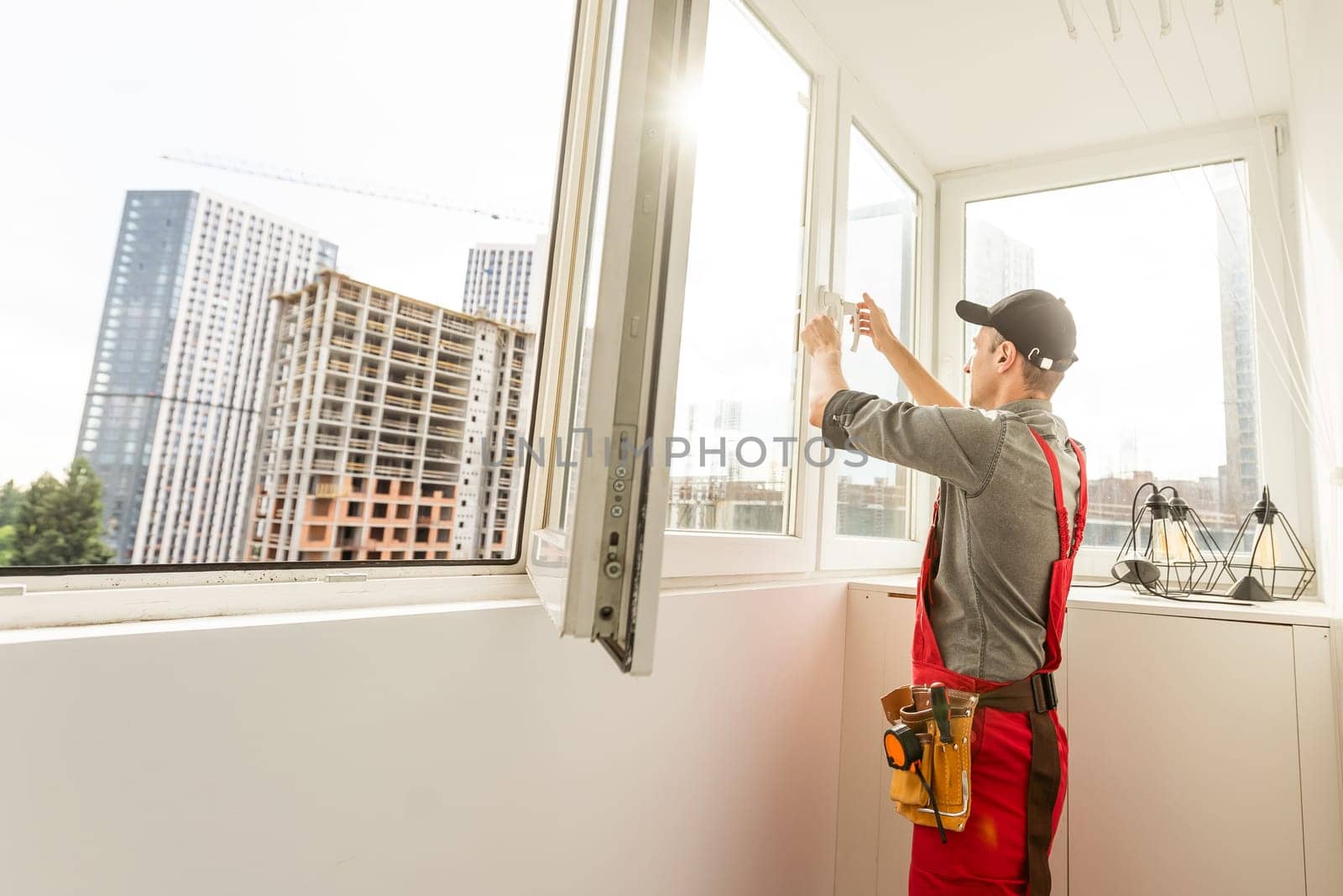 Maintenance man is fixing windows in a living room using a skrewdriver. He is wearing glasses, yellow gloves and a black apron. High quality photo