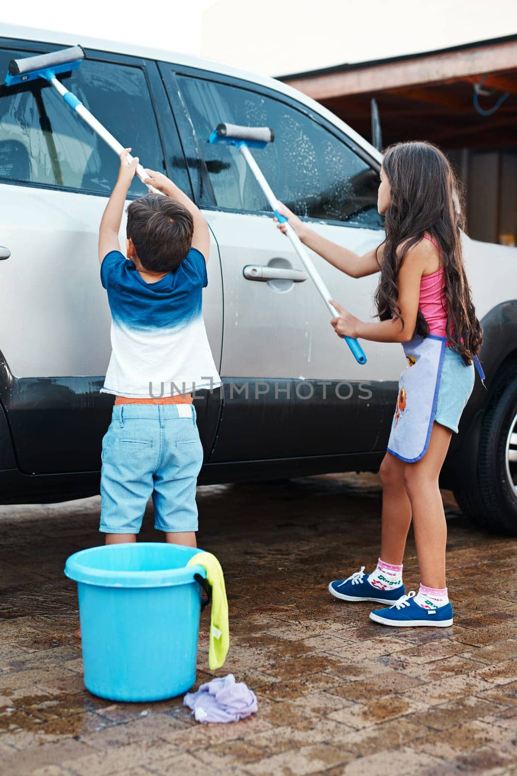 Children, clean and home or washing car, kids and motor vehicle with equipment for windows. Siblings, driveway and helping with responsibility together on weekend, outdoor for chore with bucket by YuriArcurs