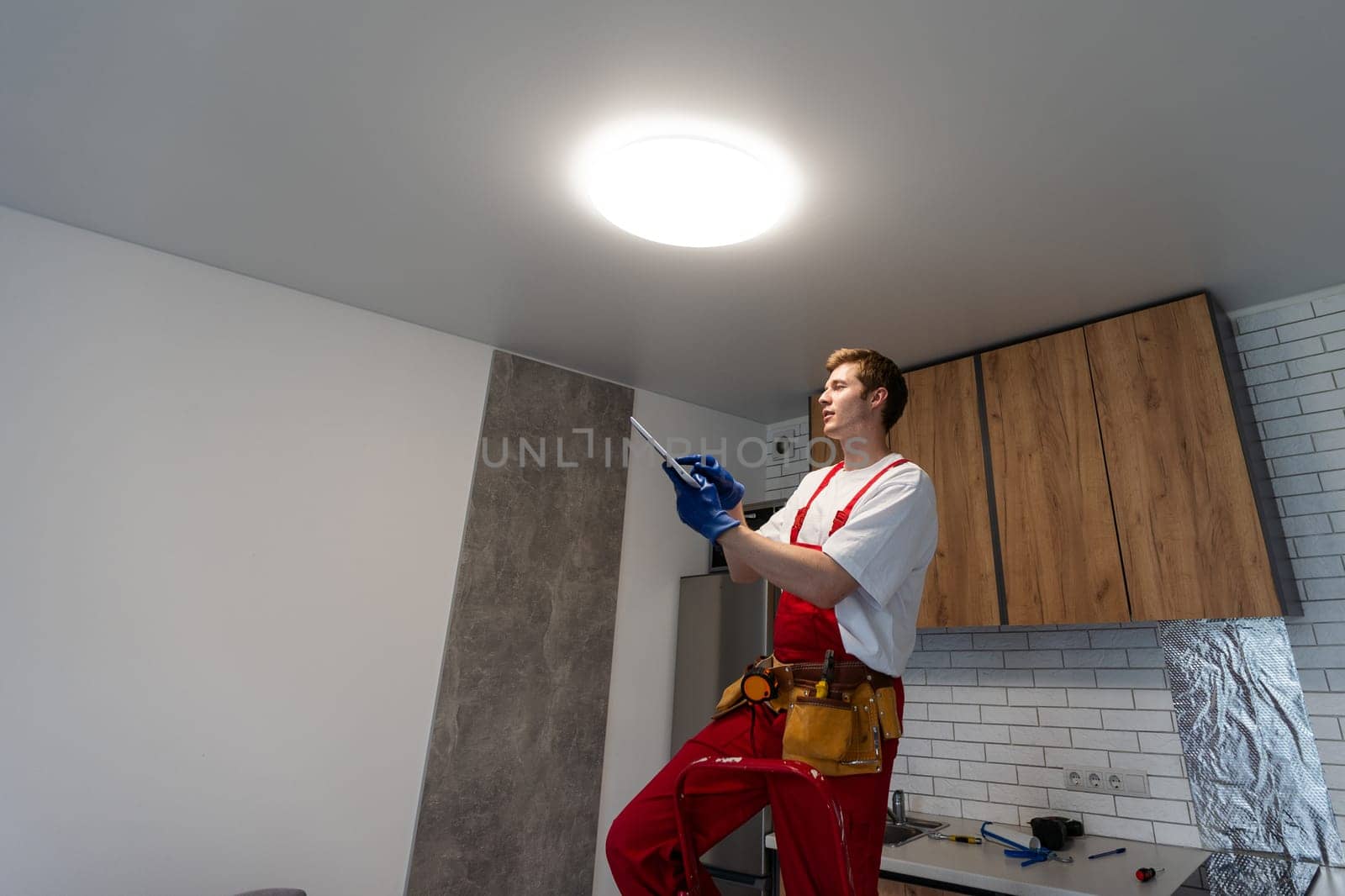 Young man installing ceiling lamp on stepladder in kitchen.