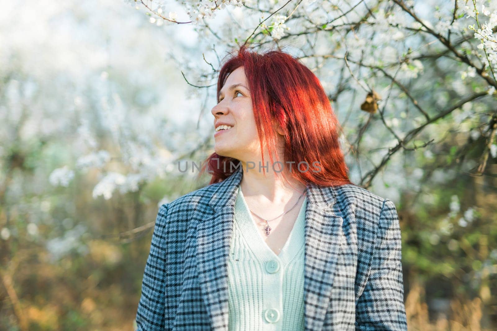 Beautiful red-haired woman enjoying smell in a flowering blooming spring garden. Spring blossom. Copy space by Satura86