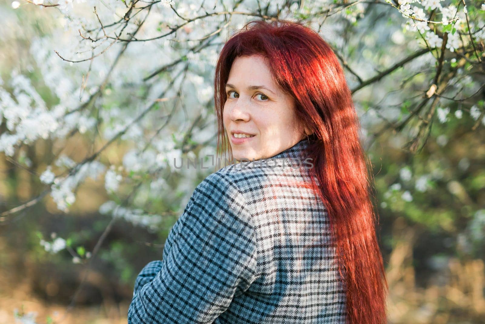 Beautiful red-haired woman enjoying smell in a flowering blooming spring garden. Spring blossom. Copy space by Satura86