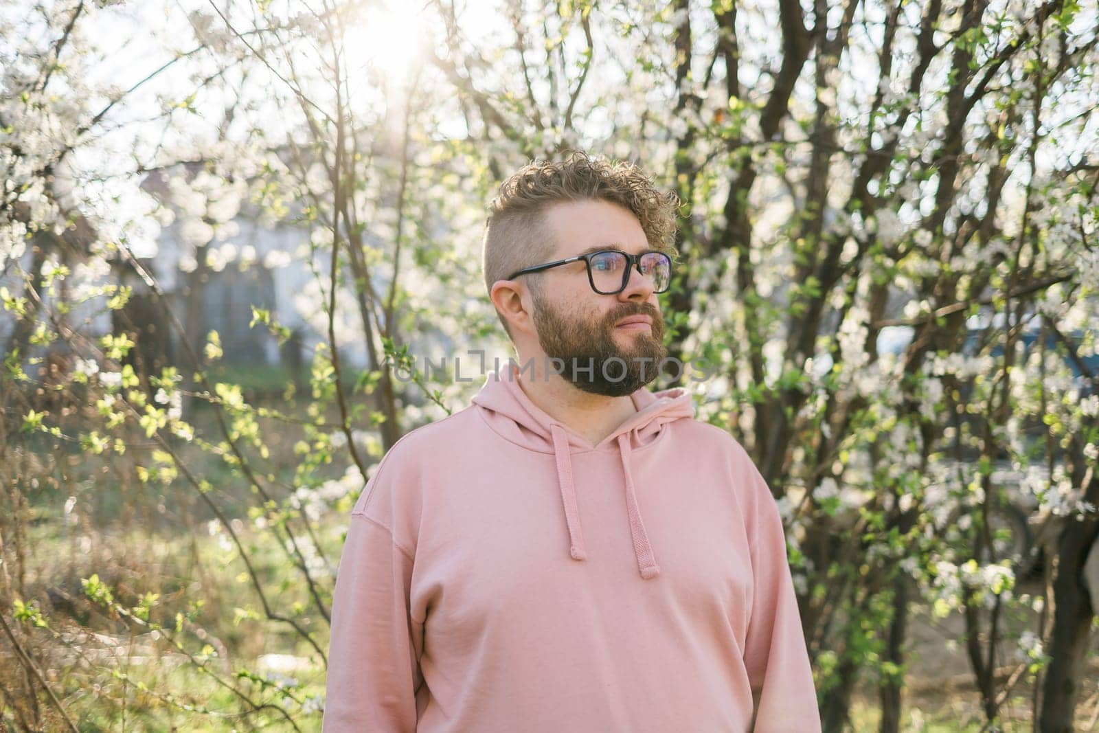 Male bearded guy standing under branches with flowers of blooming almond or cherry tree in spring garden. Spring blossom. Copy space