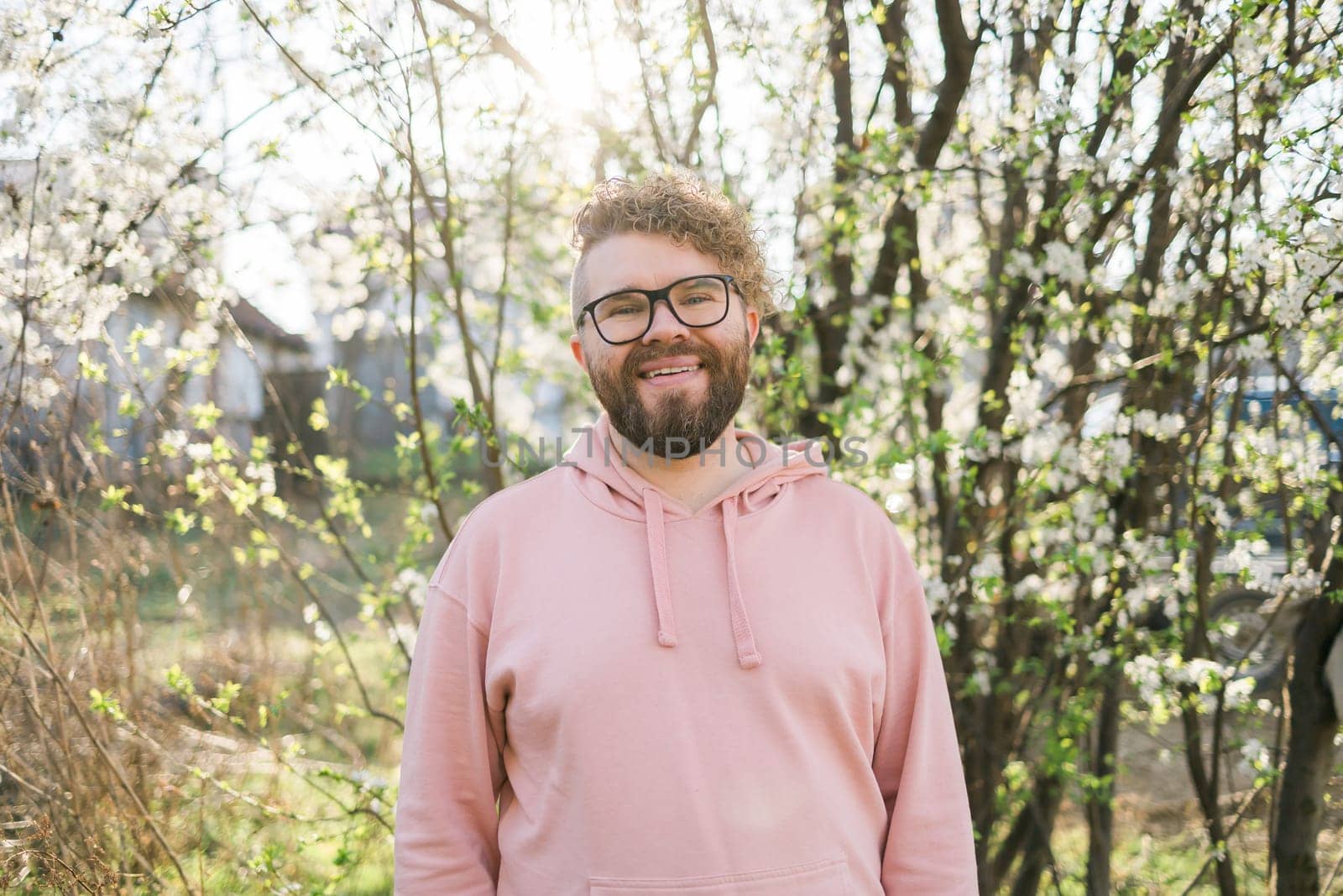 Male bearded guy standing under branches with flowers of blooming almond or cherry tree in spring garden. Spring blossom. Copy space