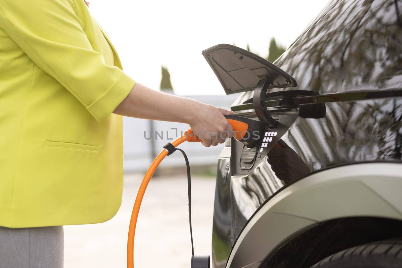 The driver of the electric car inserts the electrical connector to charge the batteries. Unrecognizable woman attaching power cable to electric car. Electric vehicle Recharging battery charging port.