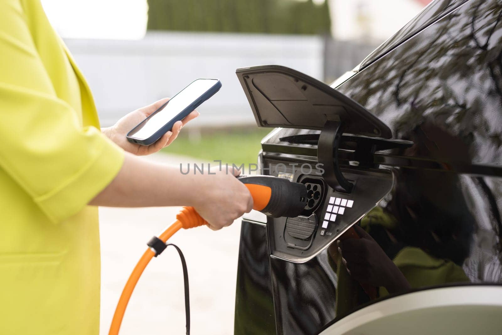 Woman unplugging electric car cable. Girl stopping charging using app on mobile phone smartphone. Eco friendly transport. Female hands disconnects plug power cable to charge electric car.