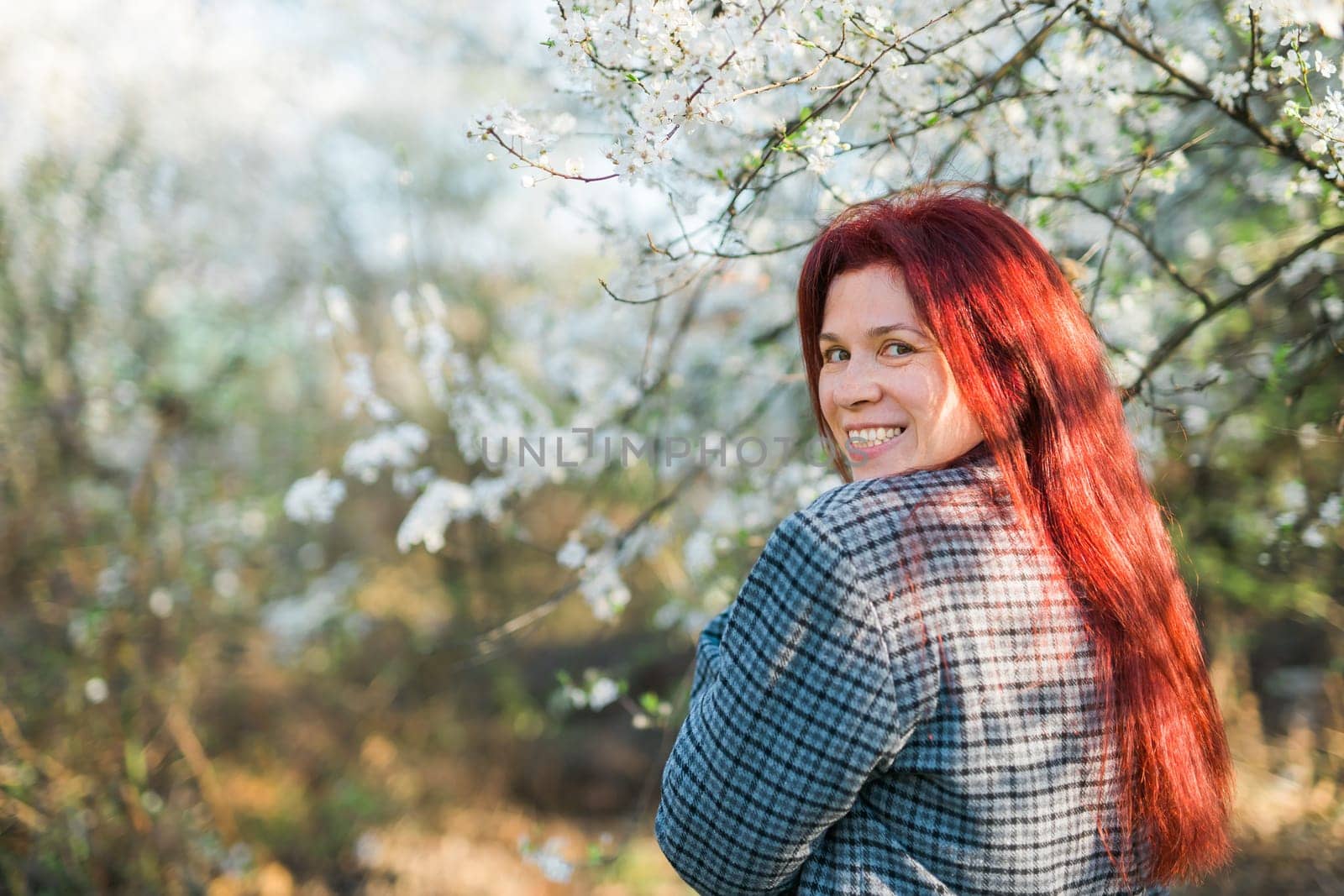 Beautiful red-haired woman enjoying smell in a flowering blooming spring garden. Spring blossom. Copy space by Satura86