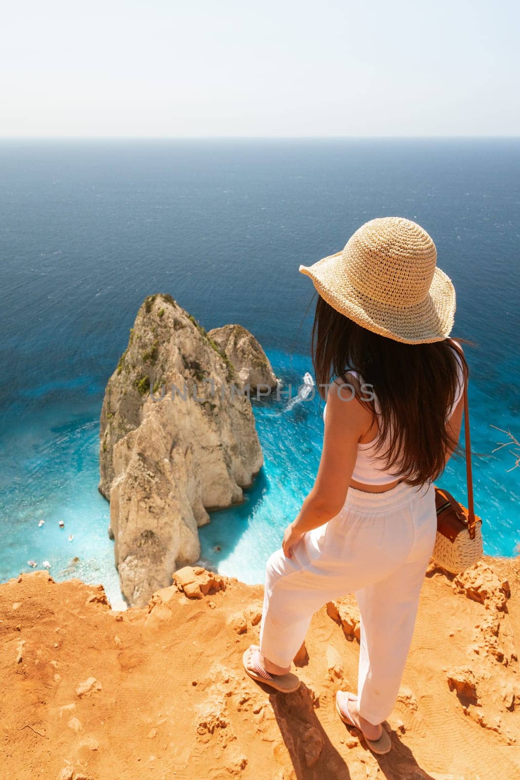 One Caucasian brunette girl in white clothes and a straw hat stands with her back on the edge of the mountain and looks at a rock in the blue sea on a sunny summer day, close-up view from above.