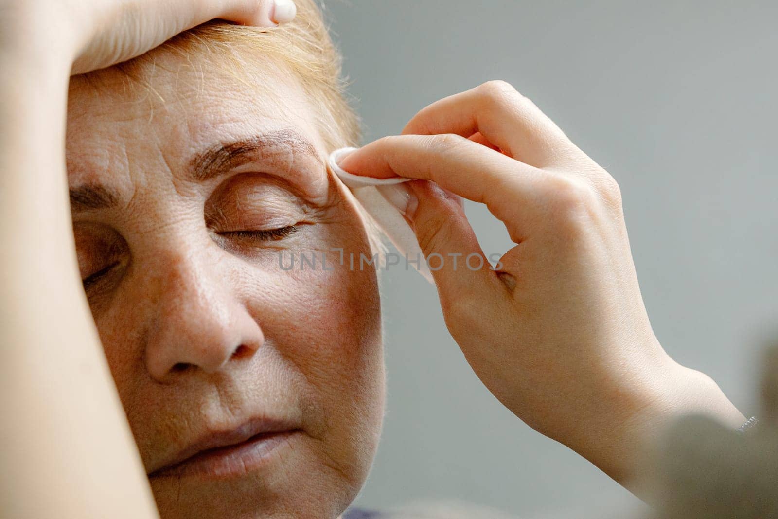 One young unrecognizable Caucasian cosmetologist wipes a tinted eyebrow with brown dye using a cotton pad to a beautiful elderly woman with closed eyes sitting in a home beauty salon, close-up side view.