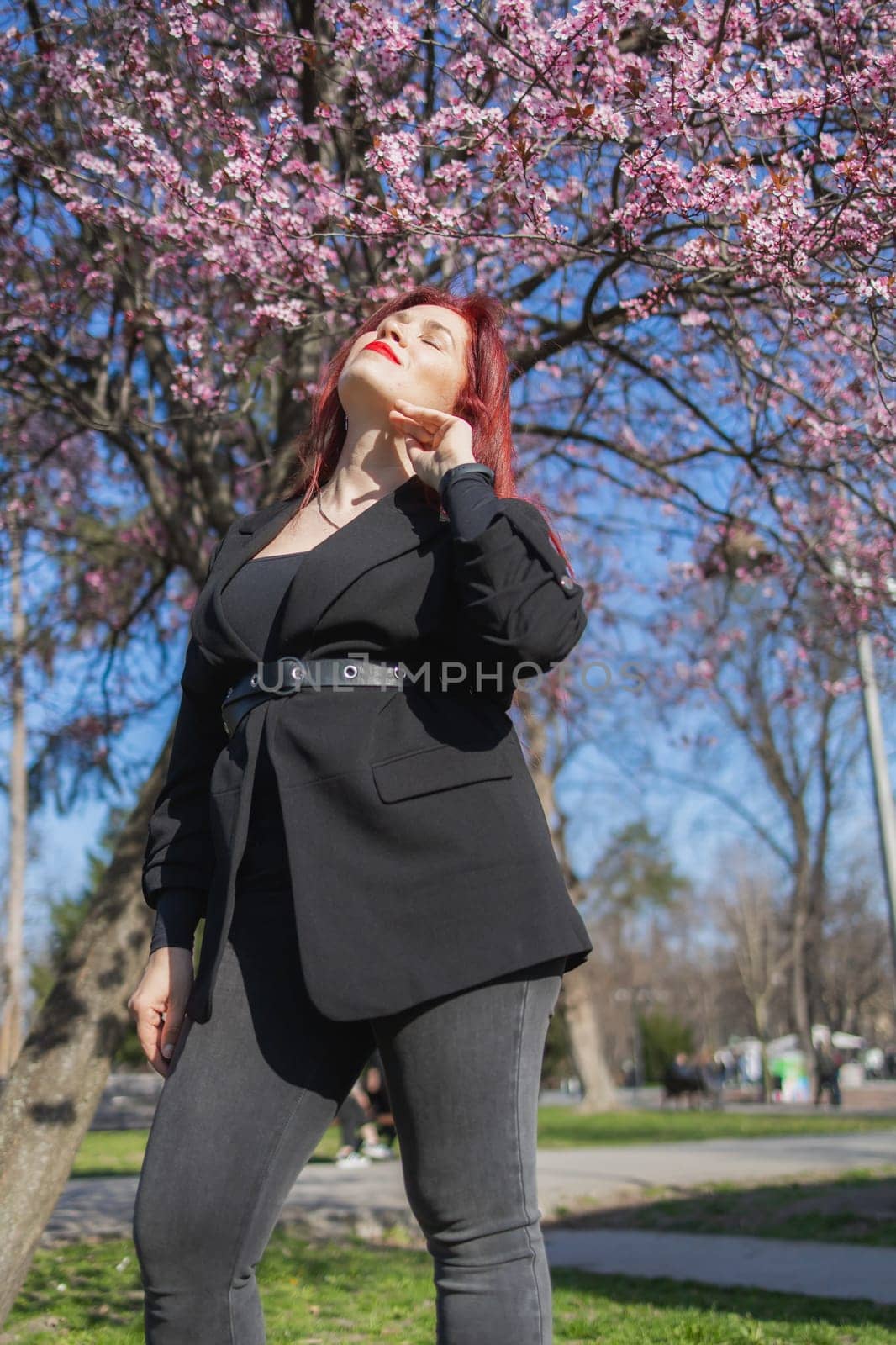 Red haired woman wearing stylish outfit near blossoming sakura in park. Fashionable spring look. Springtime blooming female portrait. by Satura86