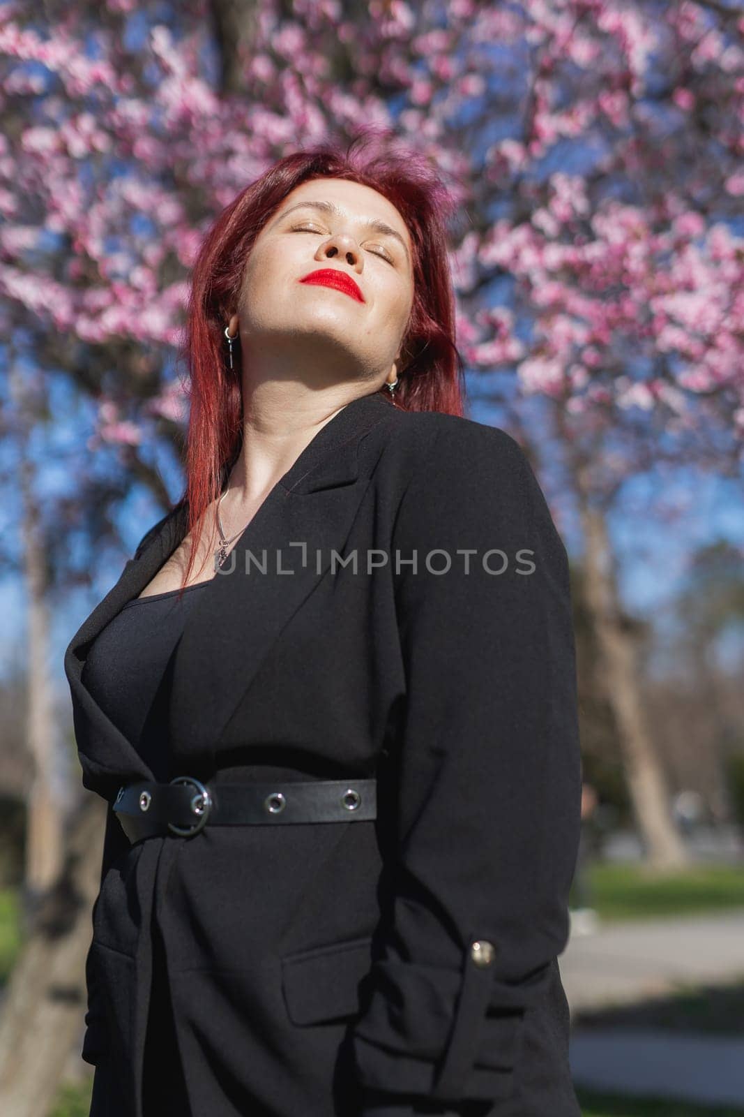 Red haired woman wearing stylish outfit near blossoming sakura in park. Fashionable spring look. Springtime female portrait