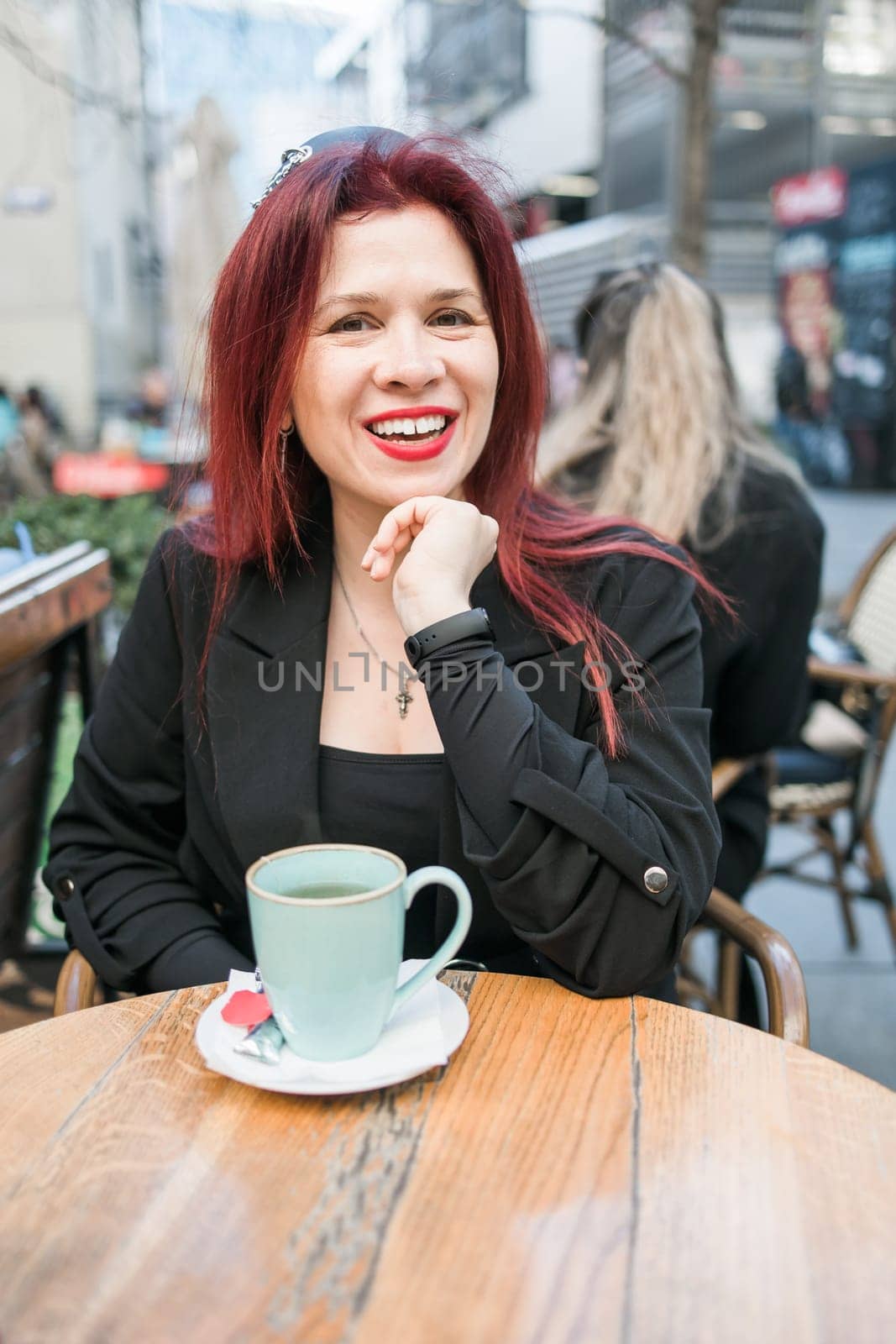 Beautiful woman with bright red lipstick sitting on restaurant terrace. Wonderful woman with red hair in black clothes posing in cafe