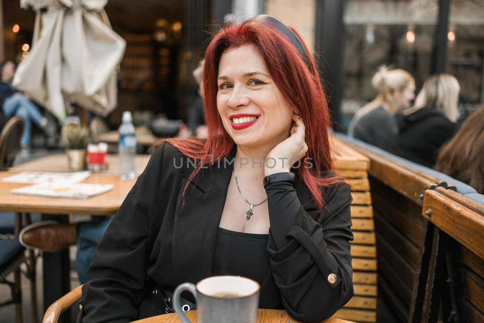 Beautiful woman with bright red lipstick sitting on restaurant terrace. Wonderful woman with red hair in black clothes posing in cafe