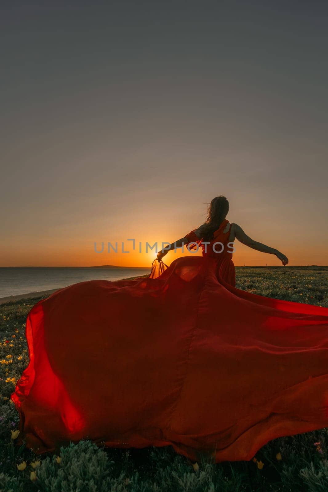 A woman in a red dress is standing in a field with the sun setting behind her. She is reaching up with her arms outstretched, as if she is trying to catch the sun. The scene is serene and peaceful