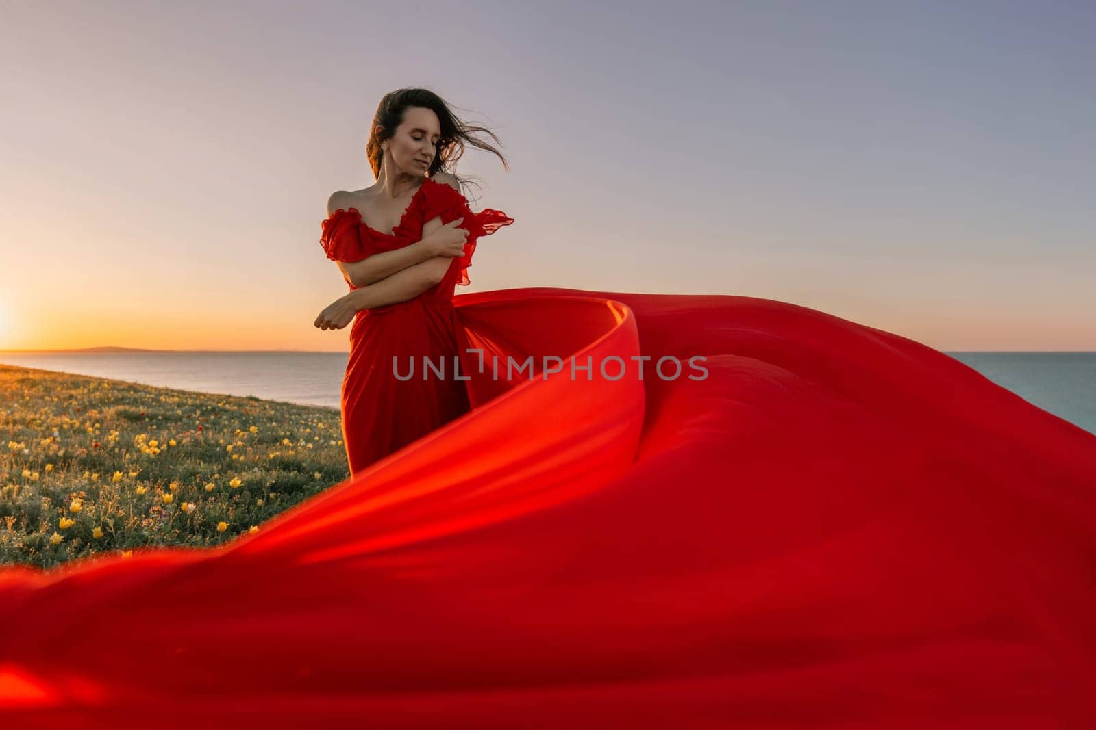 woman red dress standing grassy hillside. The sun is setting in the background, casting a warm glow over the scene. The woman is enjoying the beautiful view and the peaceful atmosphere
