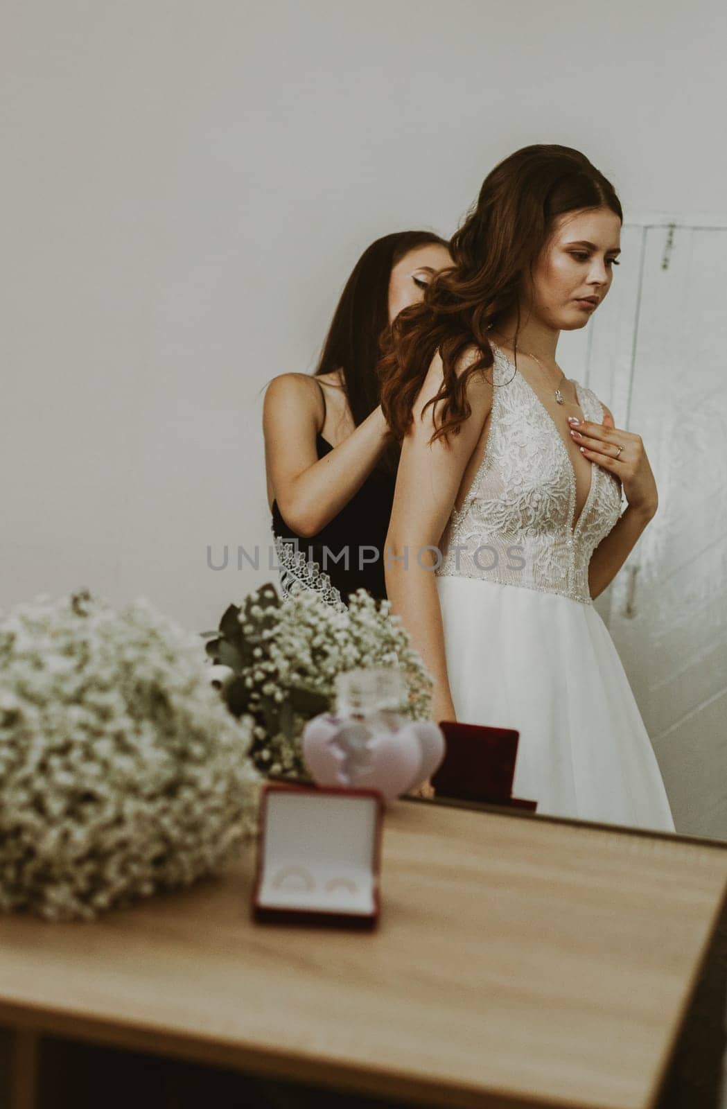 Portrait of a beautiful brunette bride and bridesmaid who fastens the clasp of a necklace around her neck, standing in a room reflected in a mirror, close-up side view.