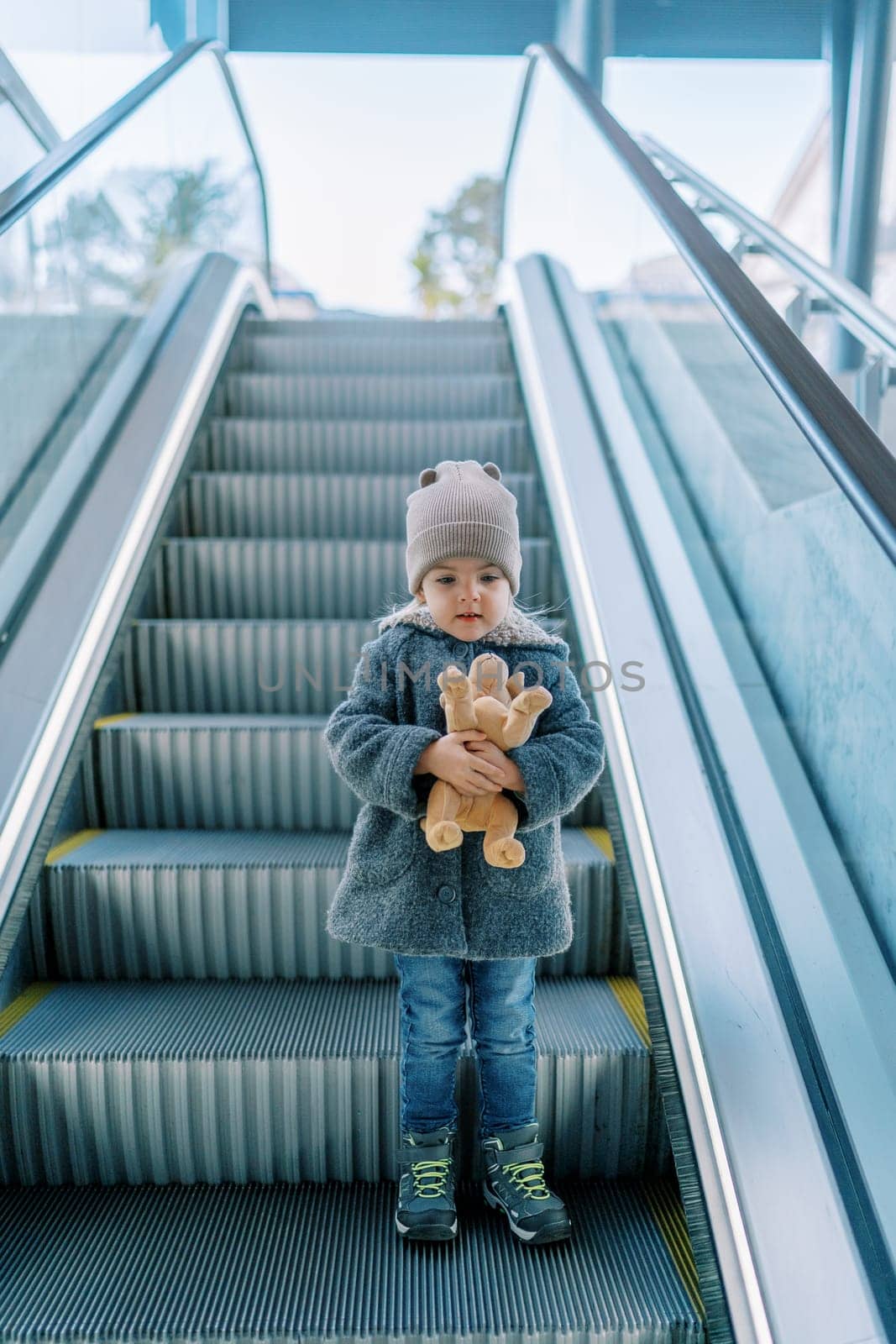 Little girl with a soft toy dog stands on an escalator and looks down. High quality photo