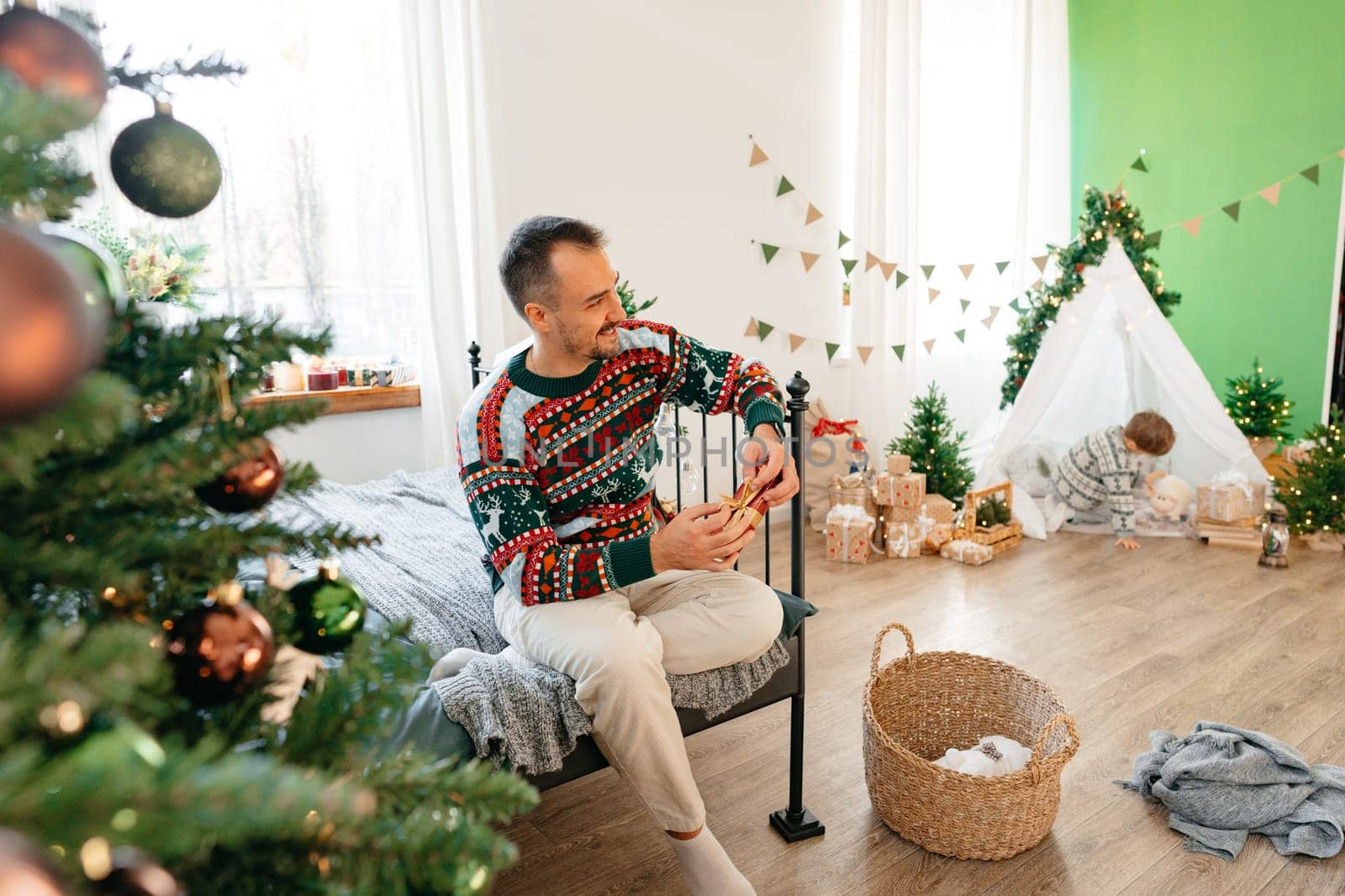 Young man in sweater sits on bed near Christmas tree at home