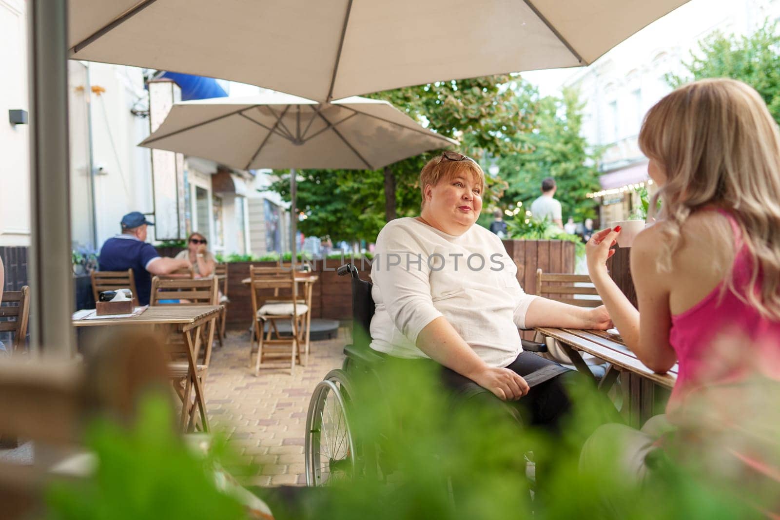 Mother with disability in wheelchair talking to her daughter while sitting at the table in cafe by Fabrikasimf