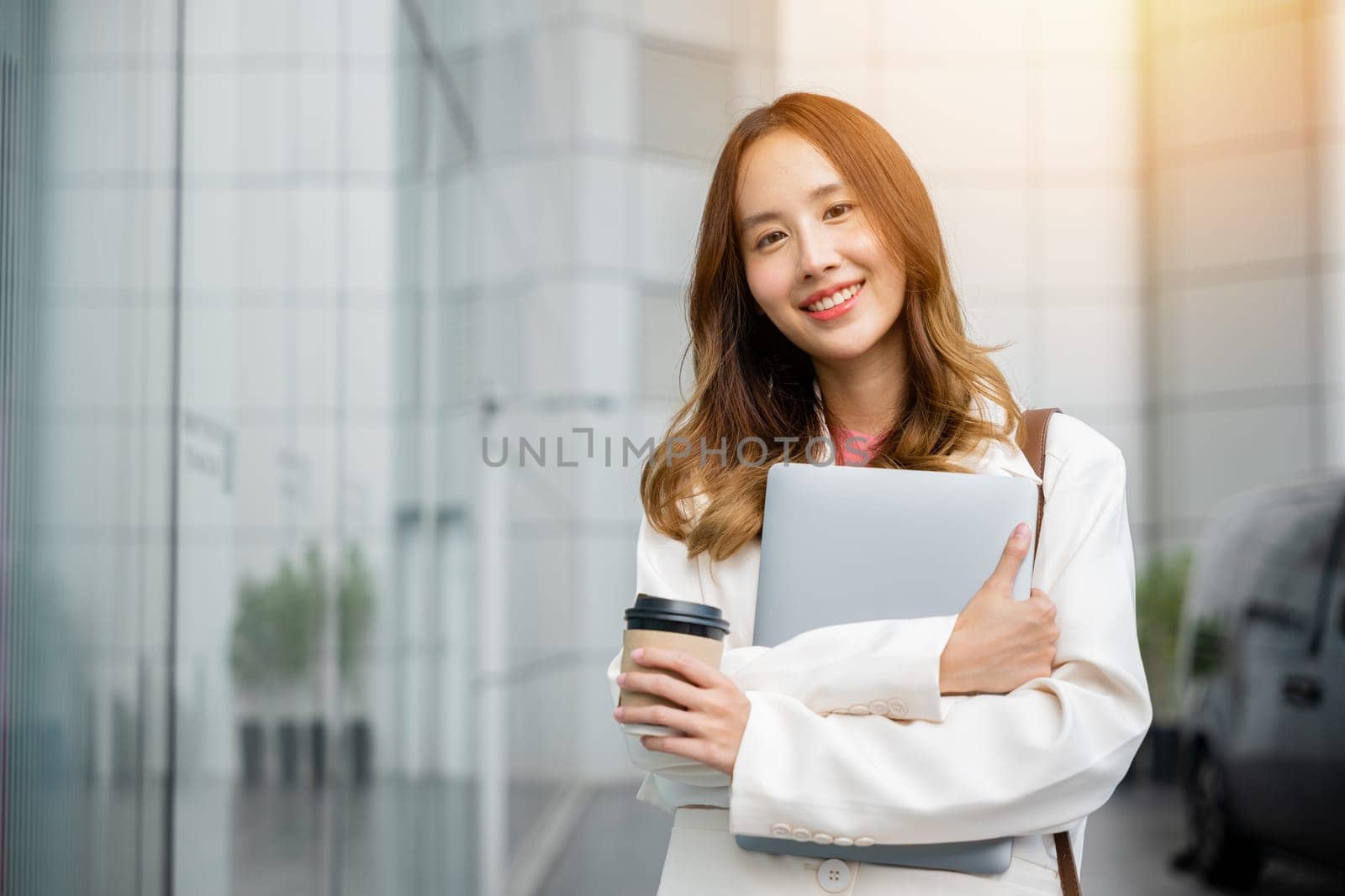 Beautiful woman enjoys a cup of coffee on go while walking outdoors with her laptop. She smiles at the camera, showing her multitasking skills and the ease of doing business online