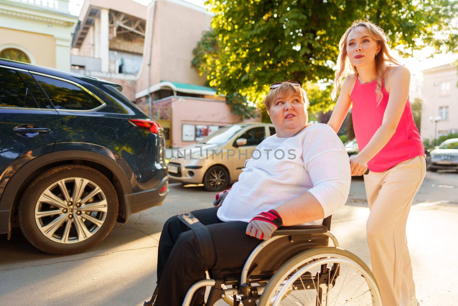 Young female caregiver pushing wheelchair with mature female person with disability across city street